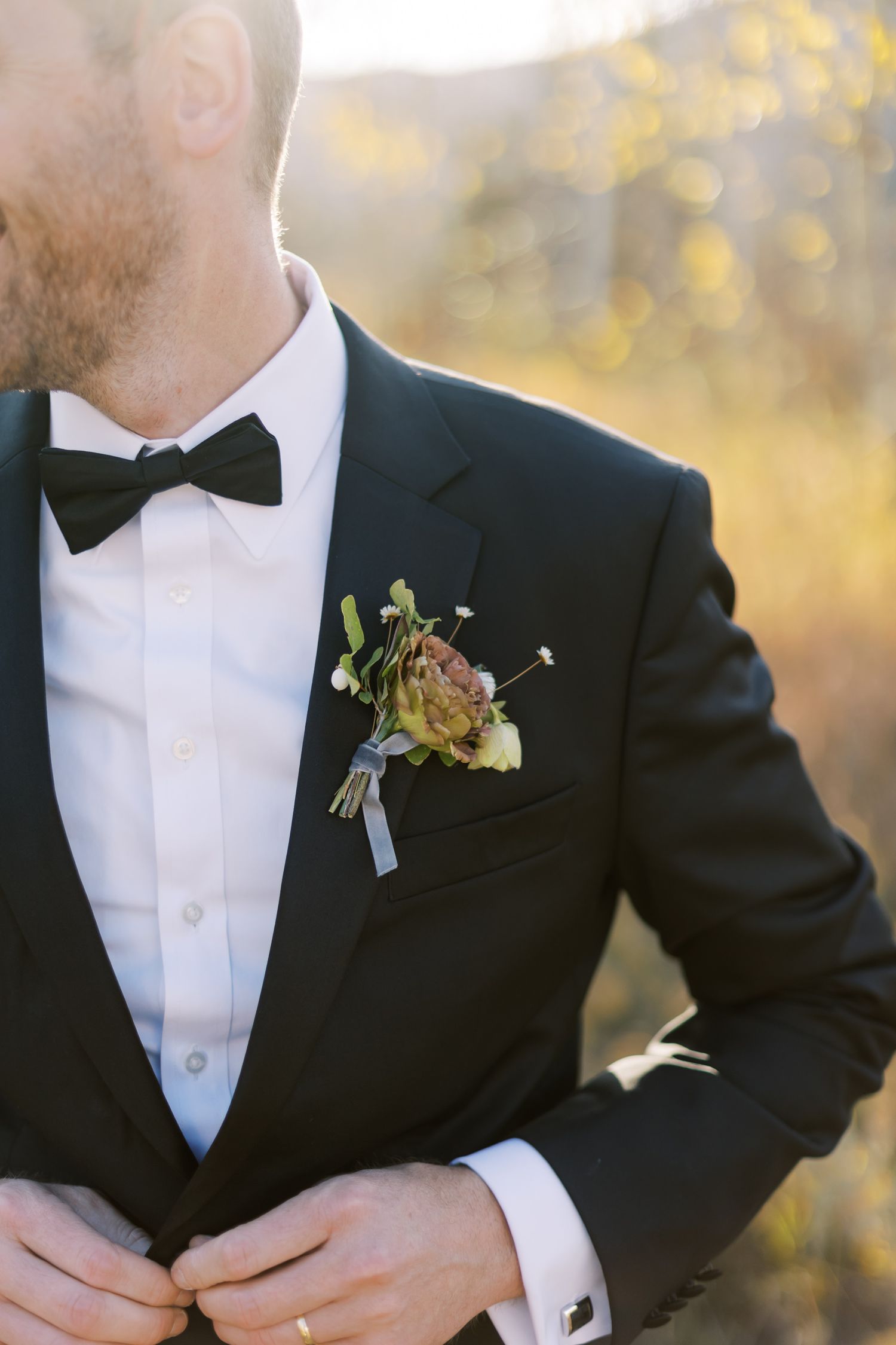 Close-up of a groom's black tuxedo with bow tie and floral boutonniere against a warm outdoor background.