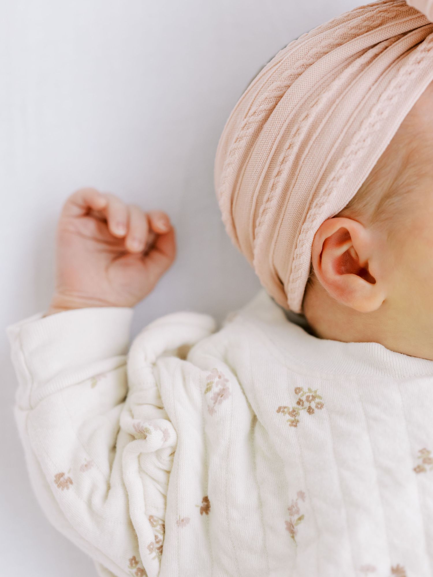 A sleeping baby in a white floral sleepsuit wearing a blush pink head wrap lies against a light background.