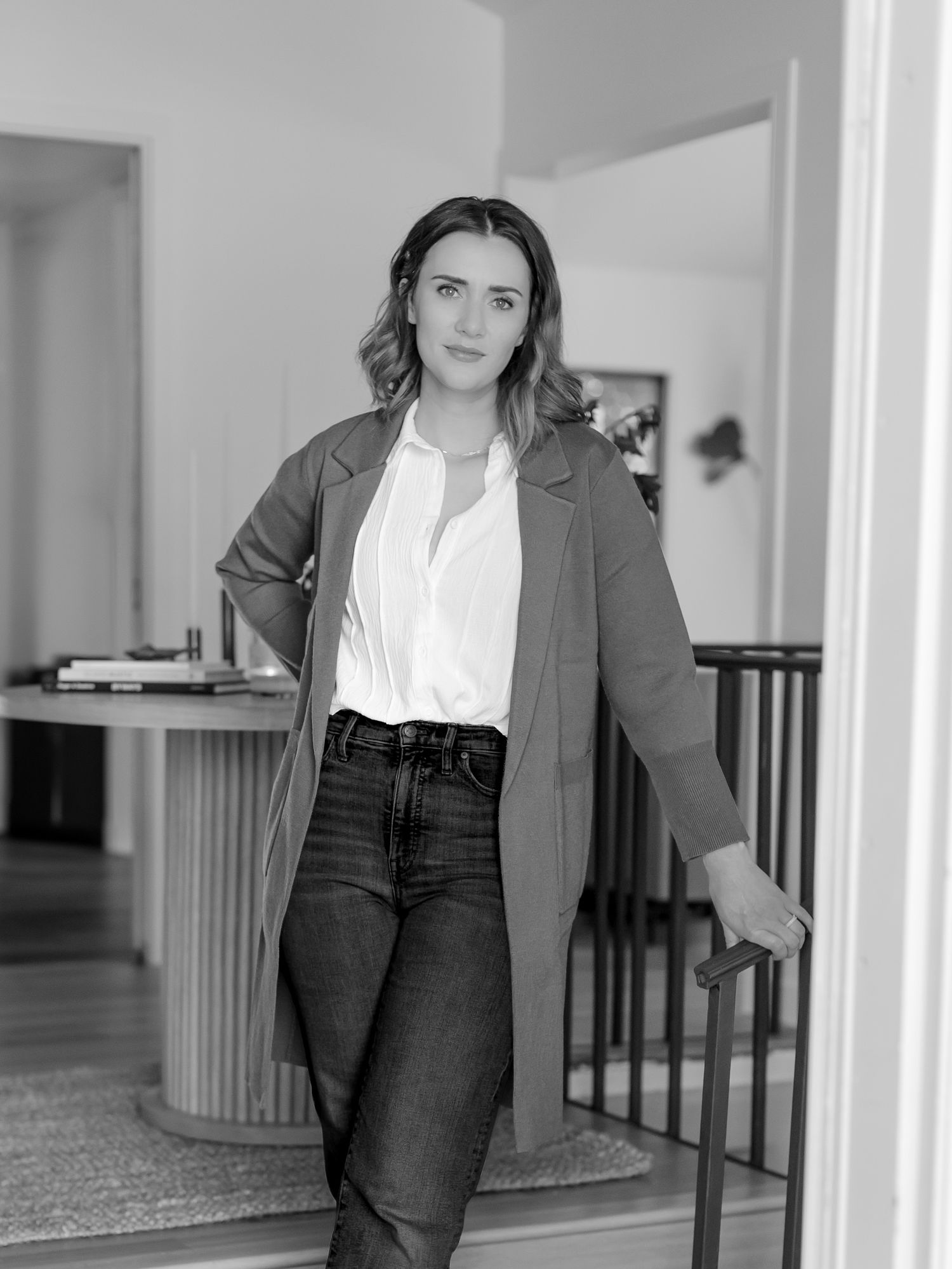 A person in a gray blazer and dark pants poses against a white wall in a modern interior hallway setting.