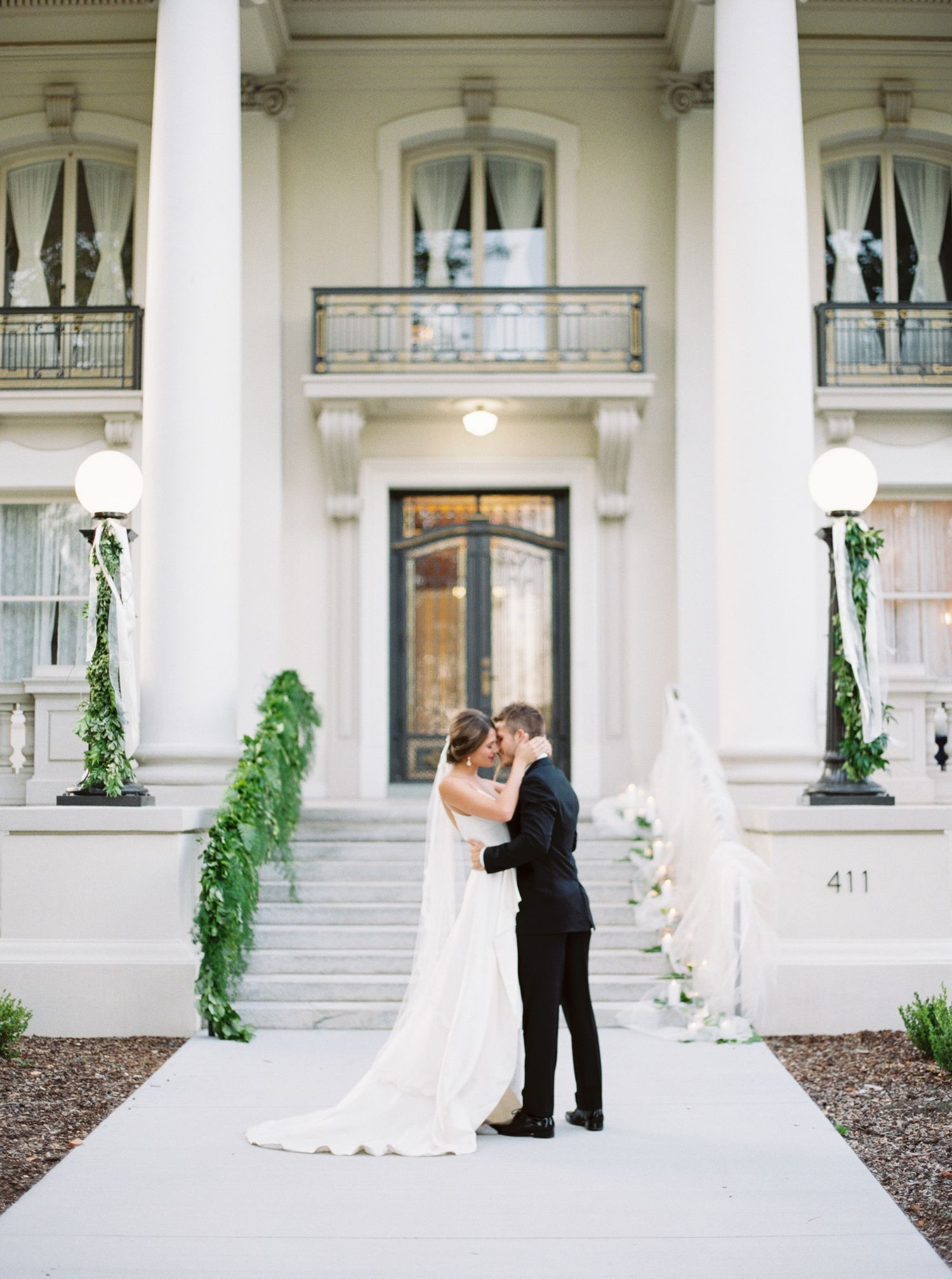 Bride and groom share a kiss in front of an elegant white mansion with columns and lush greenery at their wedding.