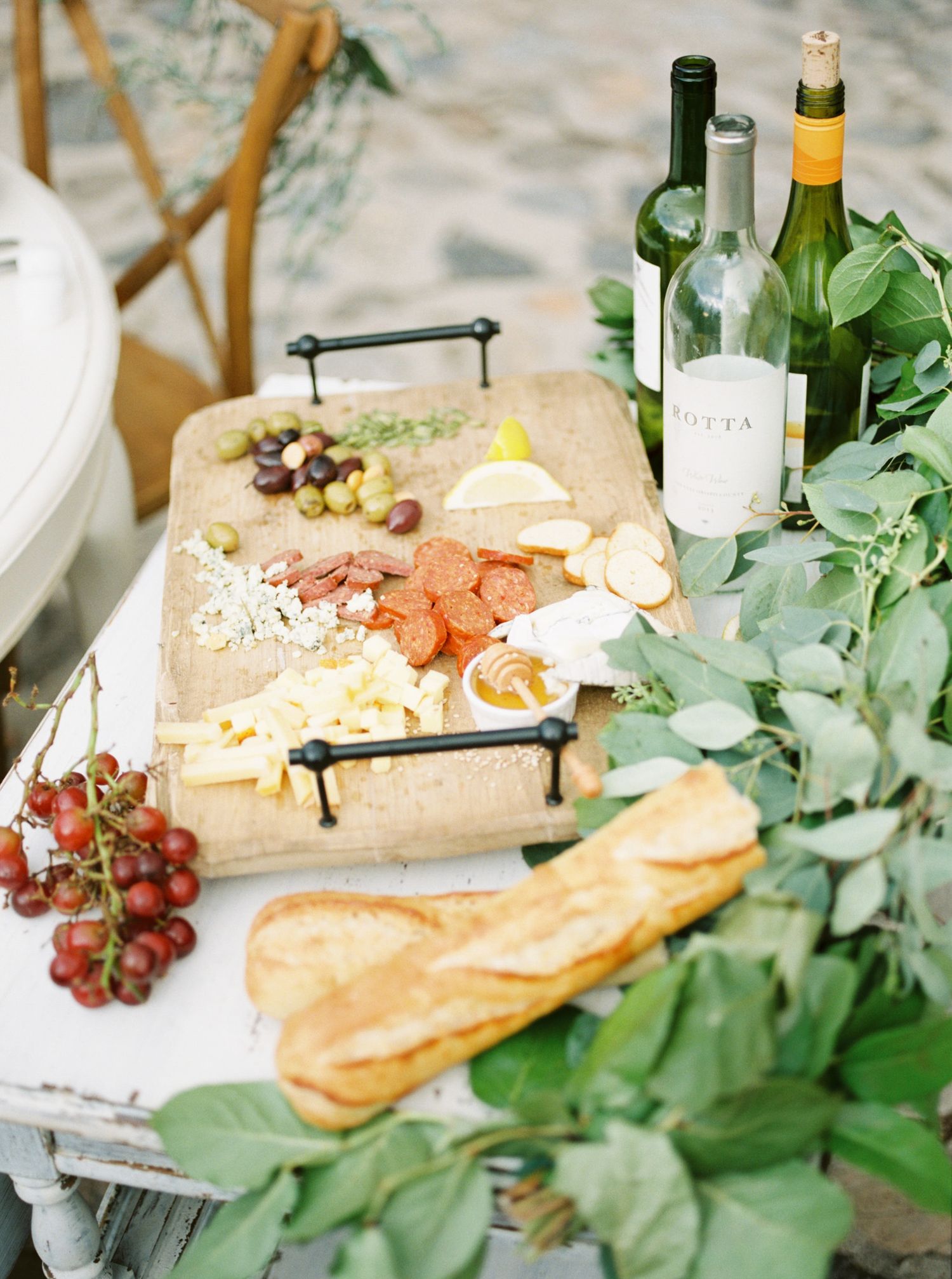 Elegant charcuterie board with cheese, meats, grapes, and honey beside wine bottles and a baguette on a white table.