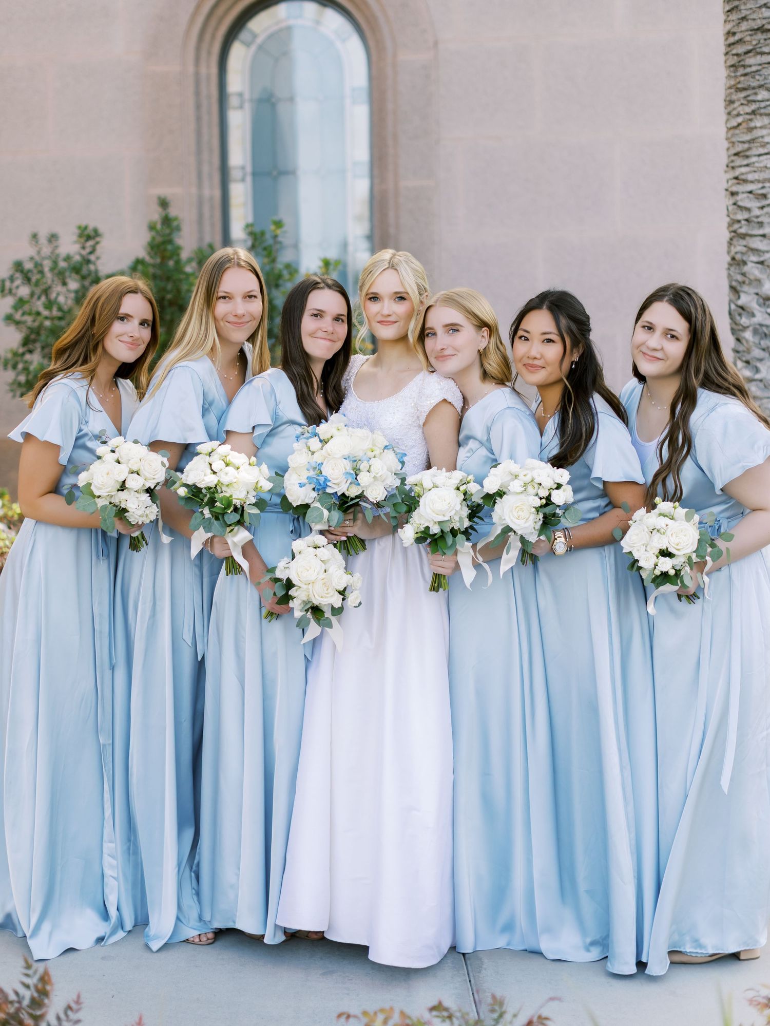 Row of bridesmaids in light blue dresses holding white floral bouquets outside a church.