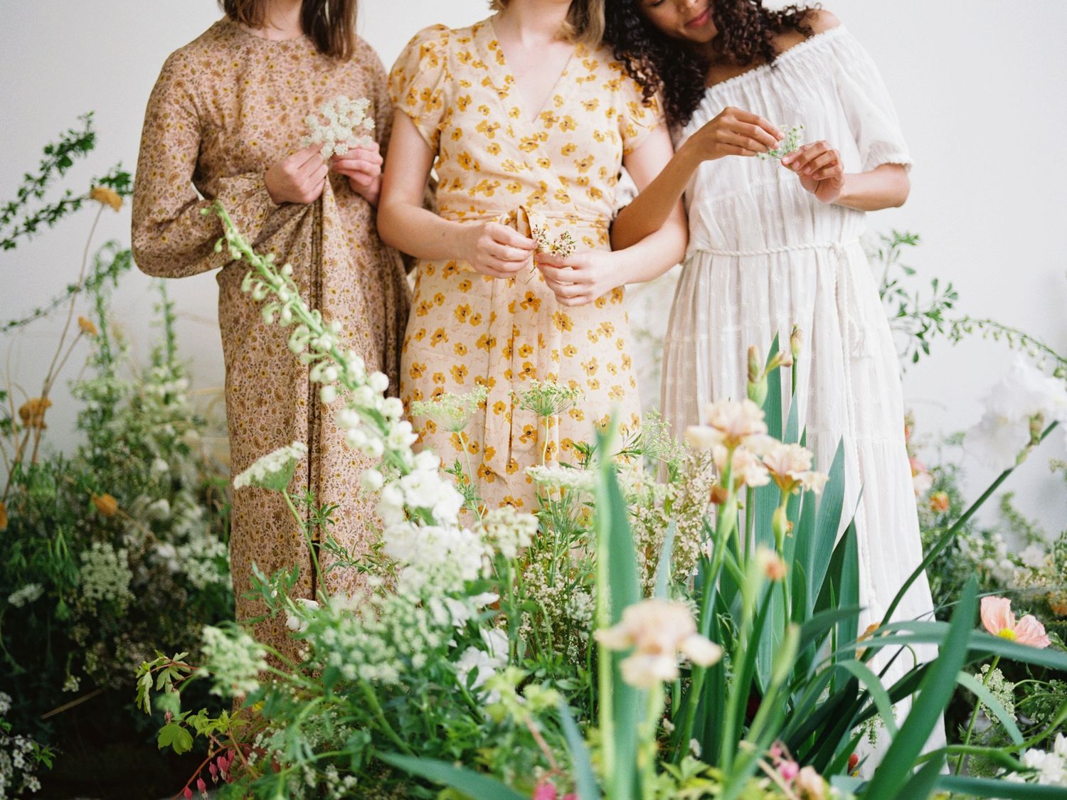 Three women in floral dresses stand among lush blooms and greenery, posing elegantly in a bright floral arrangement.