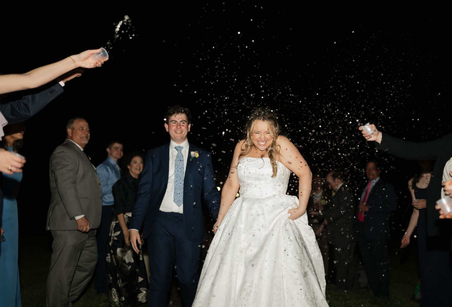 Bride and groom walk through confetti shower in New York City surrounded by cheering wedding guests at outdoor evening reception.