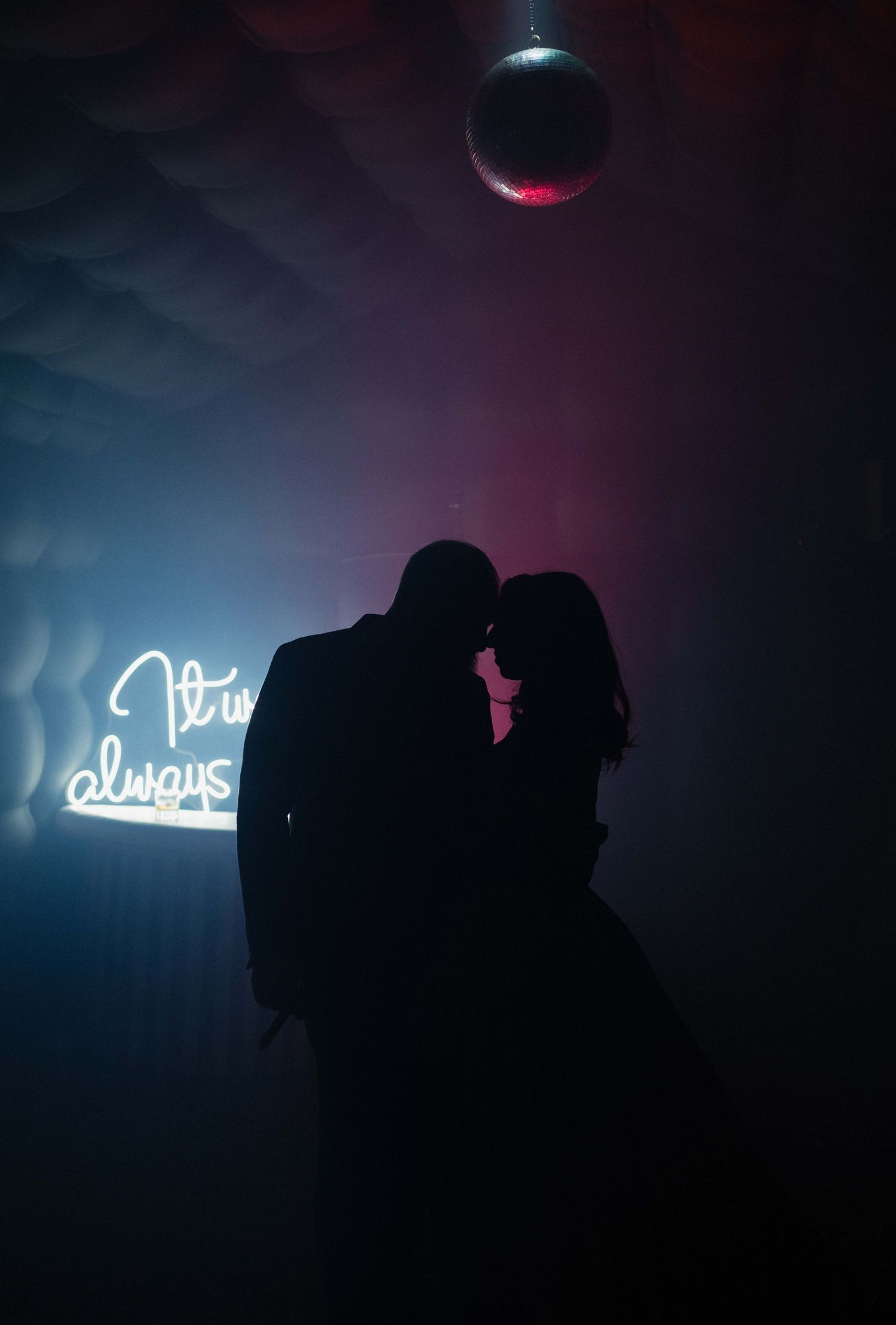 Romantic silhouette of couple dancing under disco ball in New York City with glowing neon sign reading 'It is always' in dark venue.
