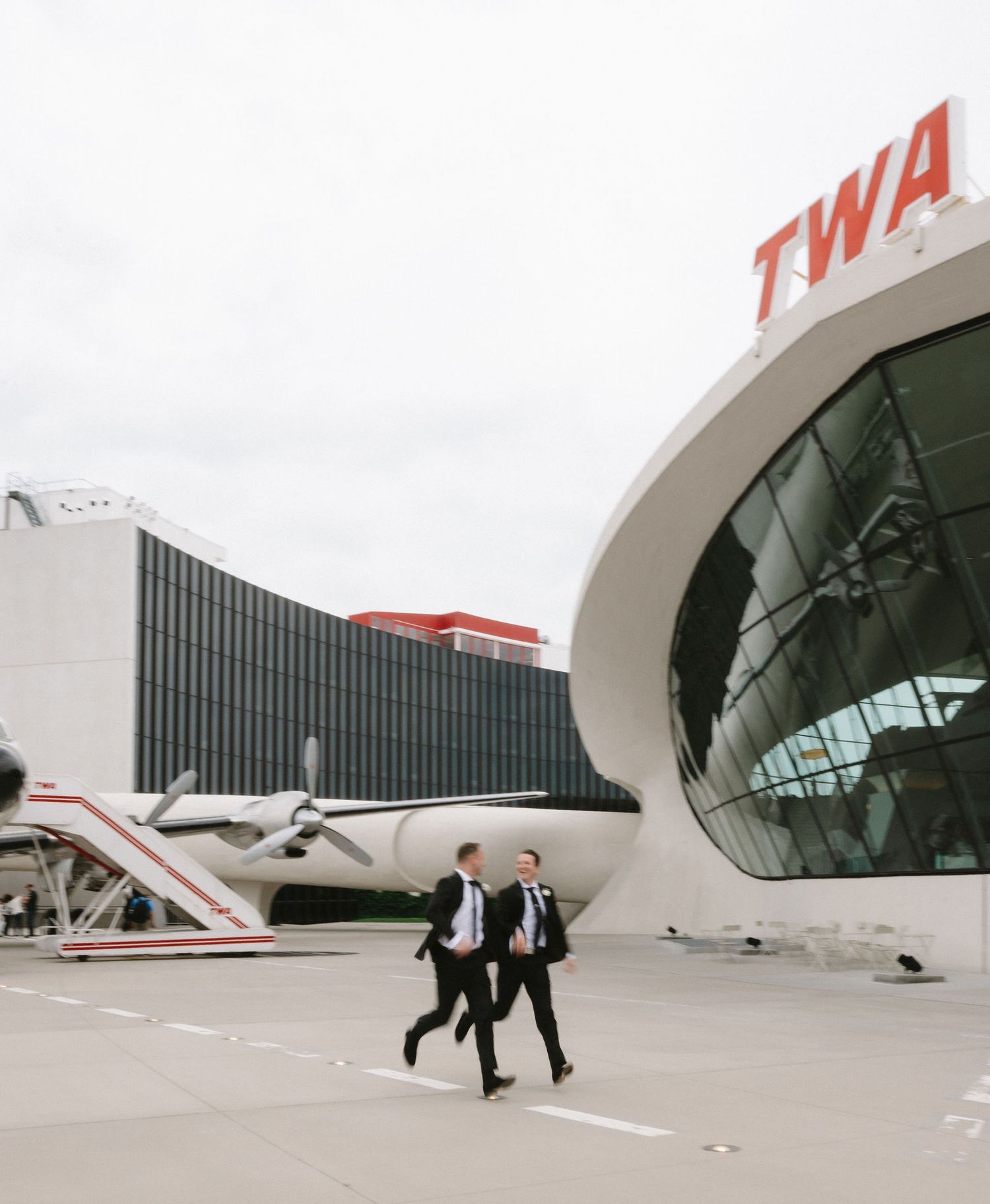 Two businessmen walking outside the iconic TWA Hotel terminal at JFK Airport with a vintage aircraft nearby.