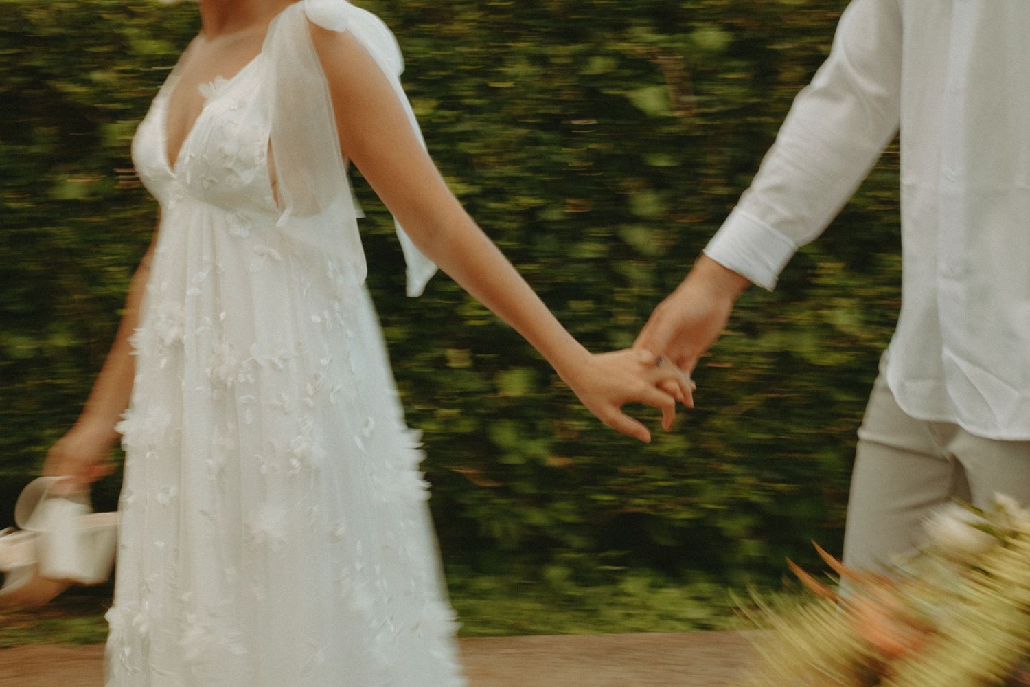 A bride in a white floral gown holds hands with a groom in white linen as they walk through a lush garden.