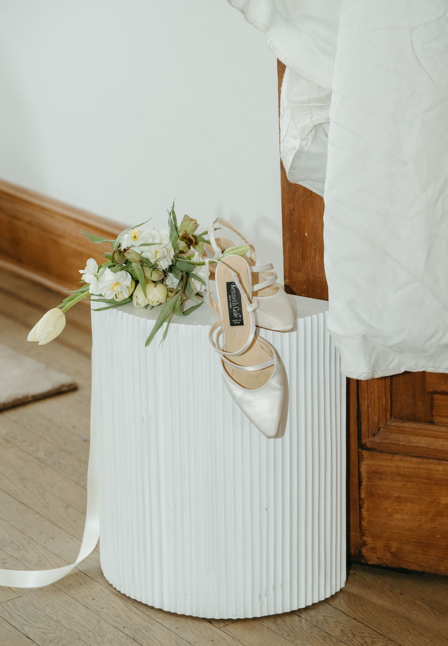 Elegant bridal heels and white floral bouquet resting on a ribbed white cylindrical pedestal near a wooden door.