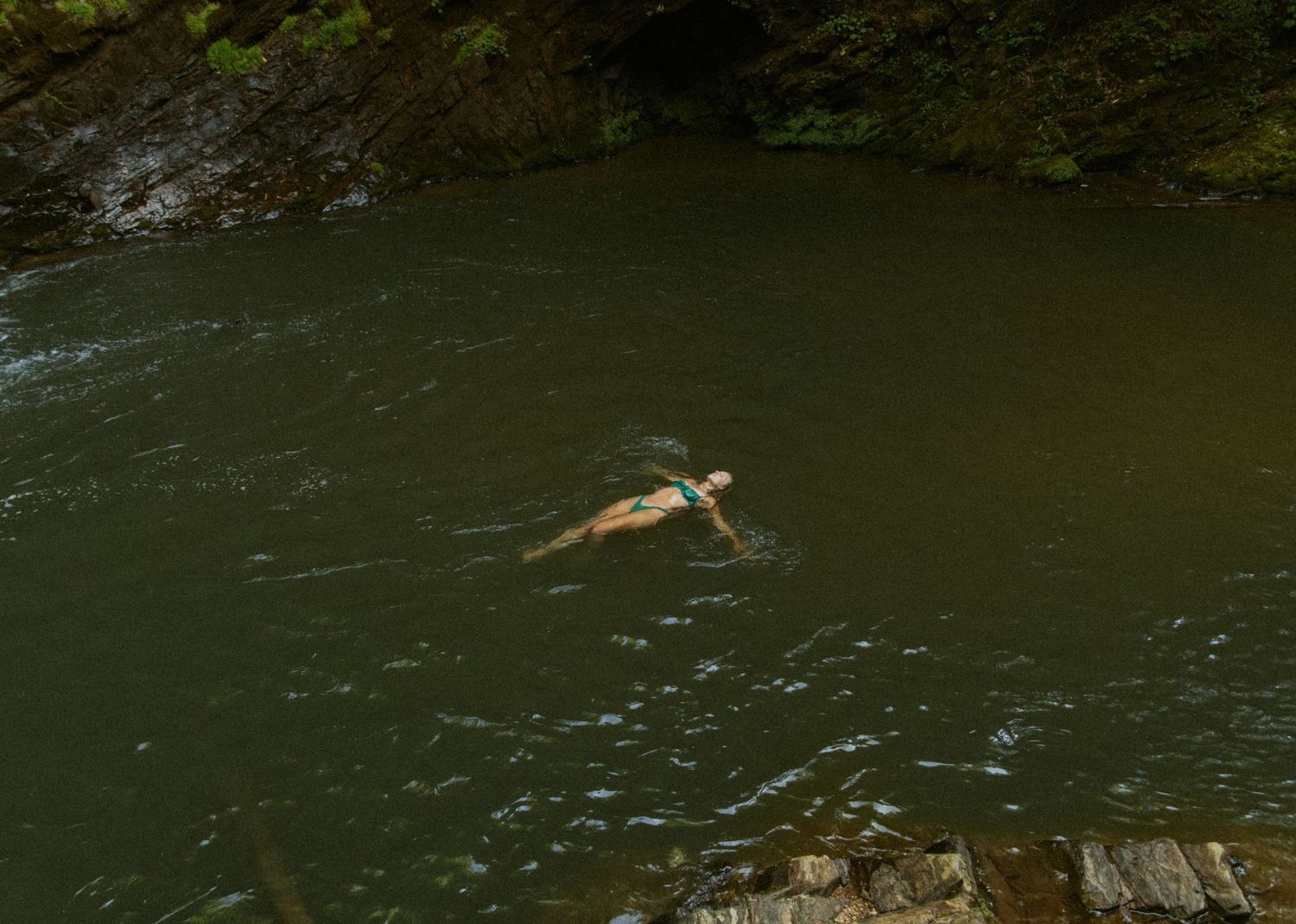 Person swimming in a serene green river surrounded by rocky cliffs and lush forest vegetation.