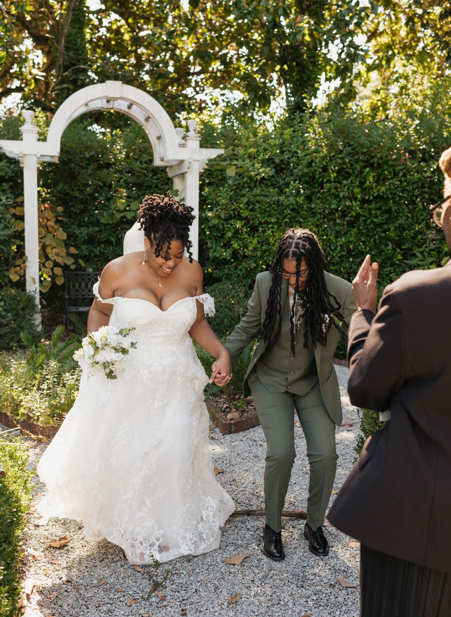 Two brides celebrate after their wedding ceremony in a lush garden, one in a white gown and one in a green suit.