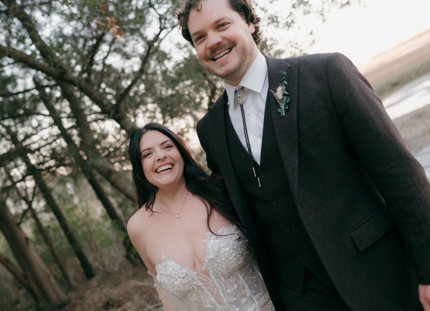 Happy bride and groom smiling outdoors in a wooded setting, wearing wedding attire on their special day.