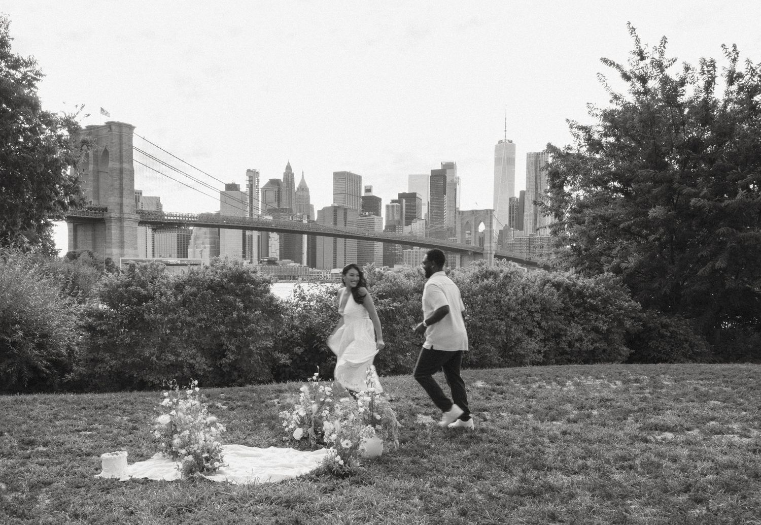 Couple walking in Brooklyn Bridge Park with NYC skyline and bridge in background, black and white wedding photo.