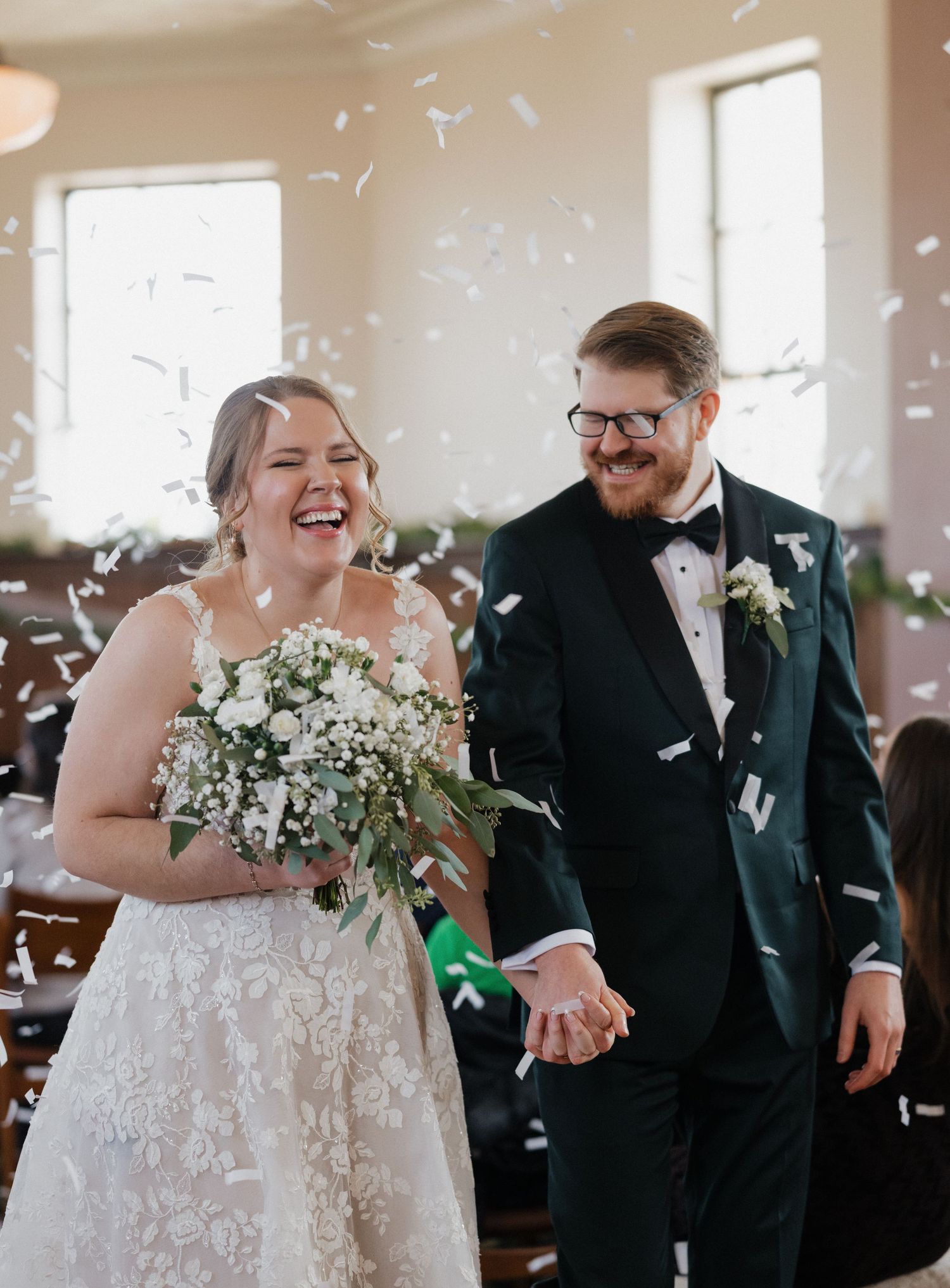 Joyful newlyweds walk down the aisle as white confetti falls, bride holding white floral bouquet in lace gown.