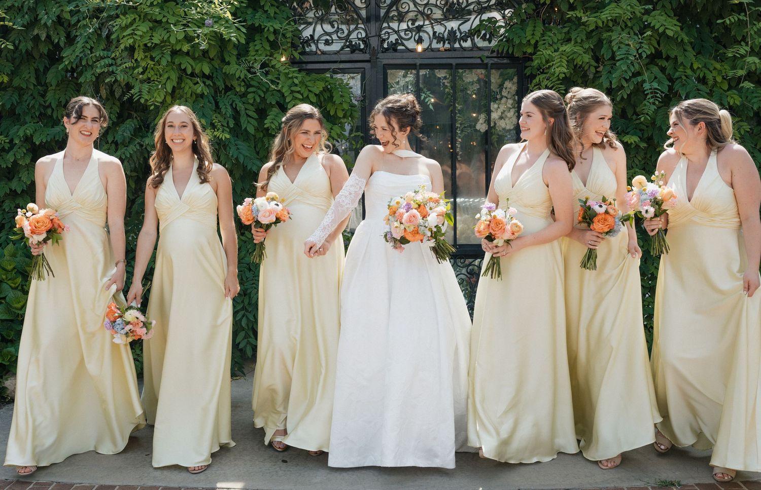 Bride in white gown walks with six bridesmaids in pale yellow dresses holding colorful bouquets outdoors.