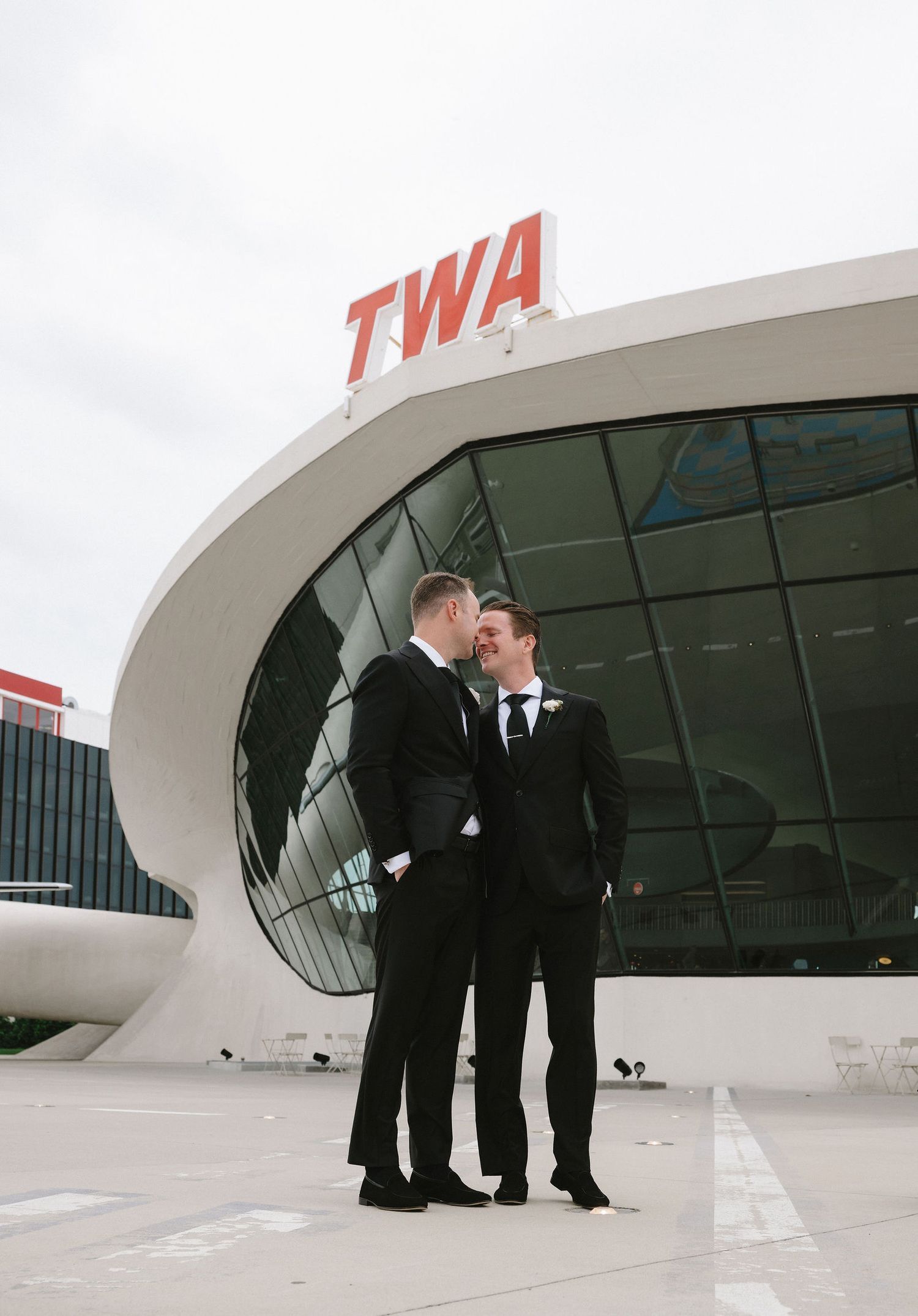 Two grooms in black suits share a romantic moment outside the iconic TWA Hotel at JFK Airport.