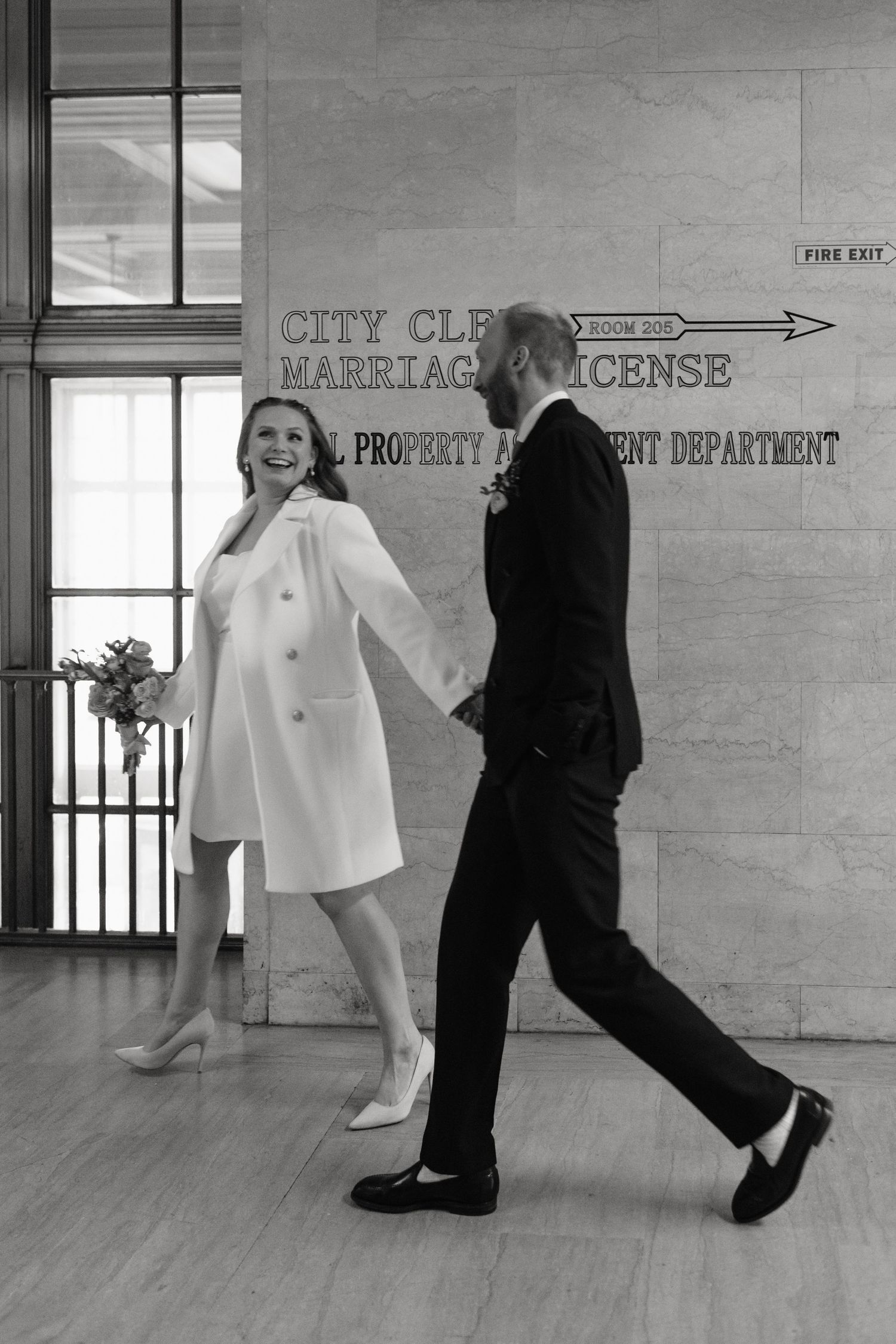 Black and white photo of a couple holding hands walking past a New York City Clerk Marriage License office in a city hall.