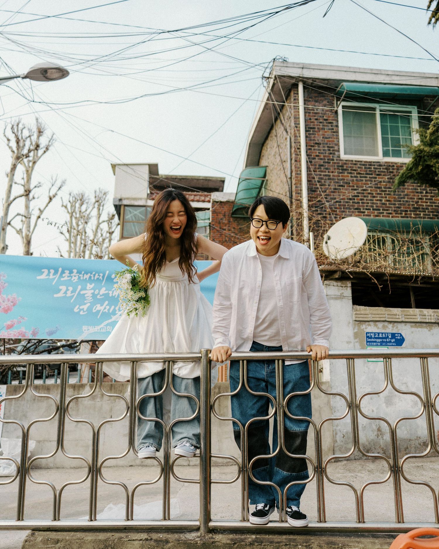 Happy couple laughing together by a metal fence in a Korean neighborhood, woman in white dress holding flowers.