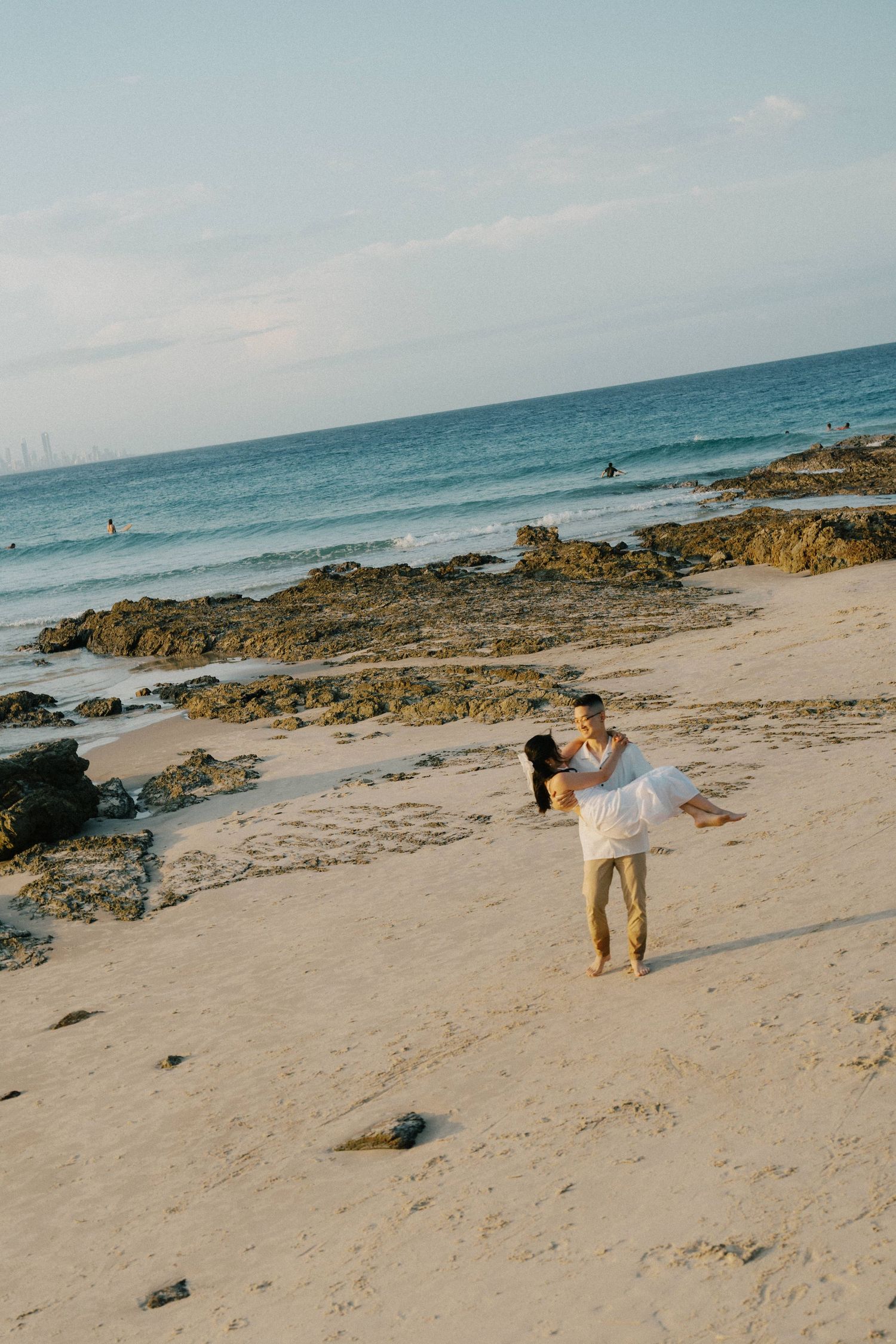 Couple embracing on a sandy beach with rocky shoreline and blue ocean waves in the background.