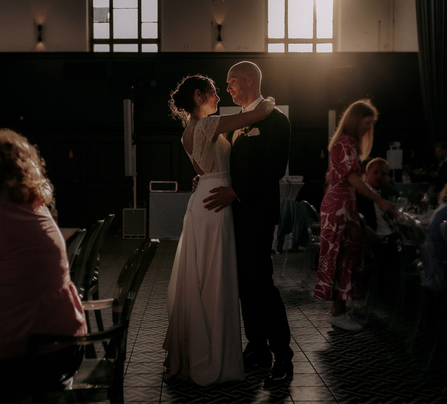 A couple shares their first dance at a dimly lit wedding reception venue with guests watching from the sides.