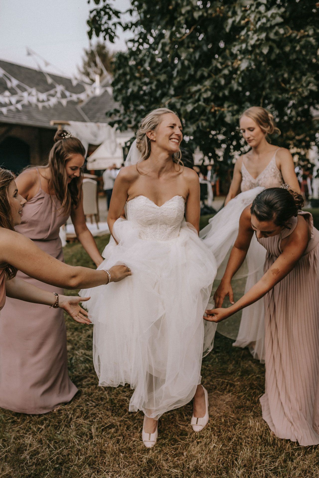 Bridesmaids in pink dresses help adjust the bride's wedding gown outdoors.