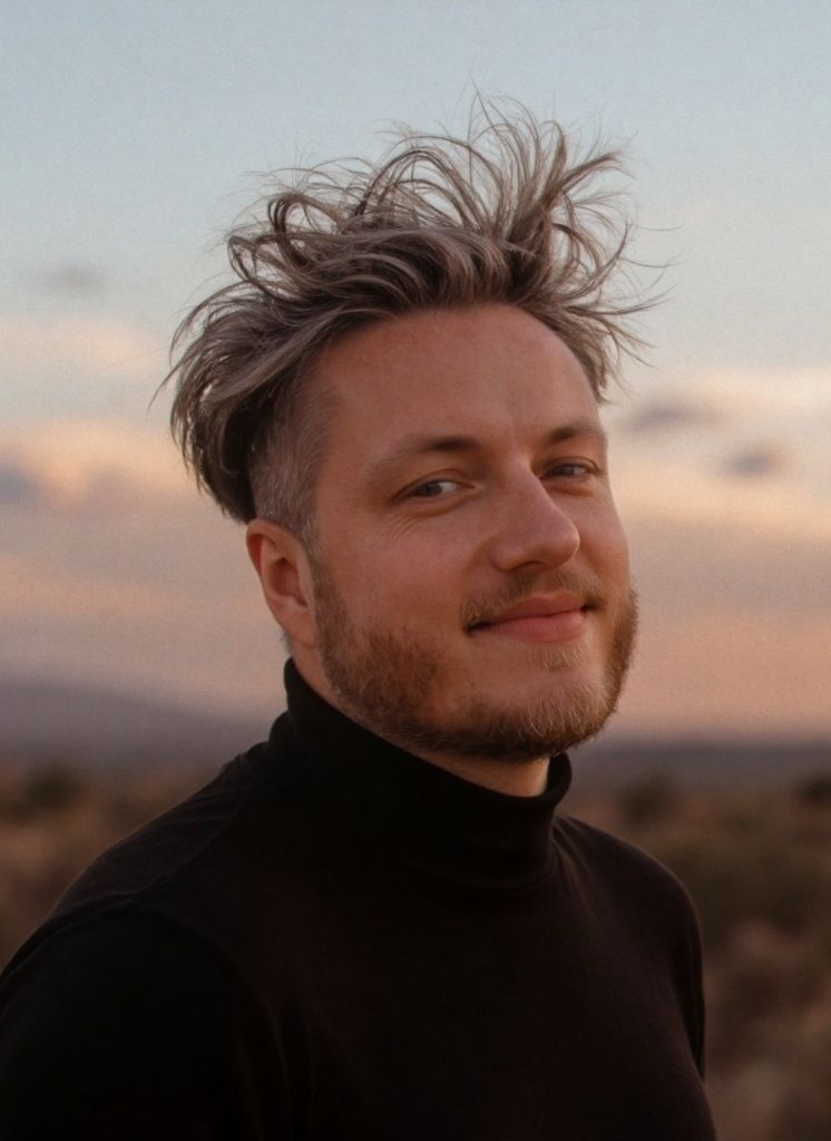Young man with blonde hair and beard wearing a black turtleneck smiling outdoors at sunset.