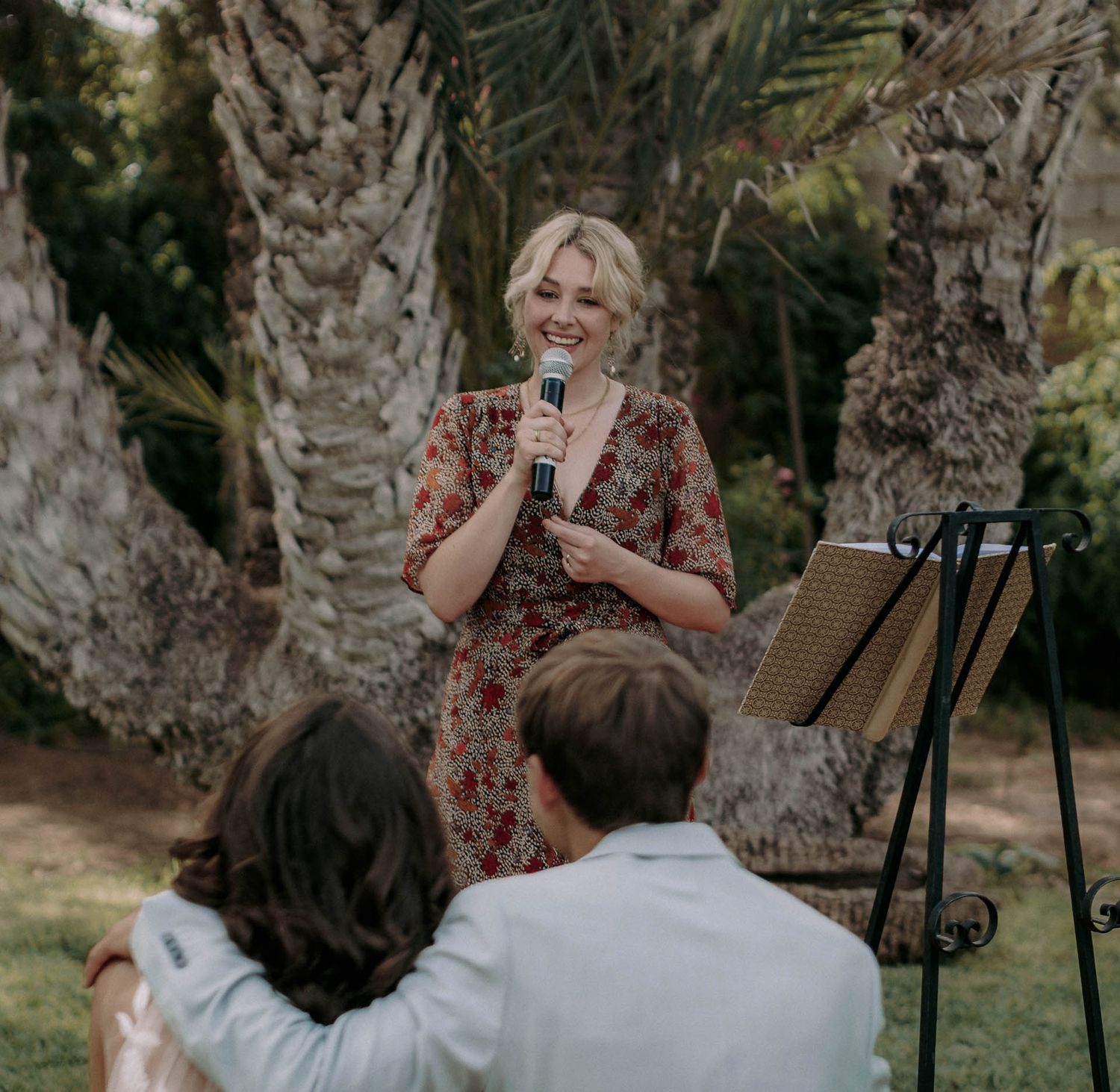 Speaker addresses wedding guests during an outdoor ceremony among palm trees.
