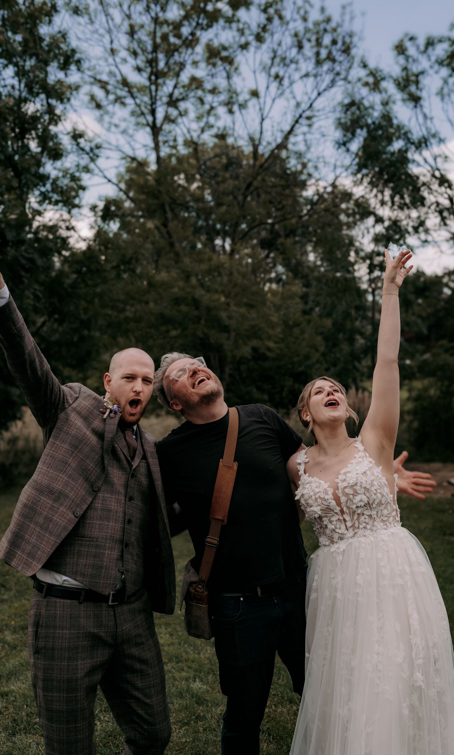 Bride in lace gown and two men in suits celebrate joyfully outdoors, arms raised, at a wedding reception.