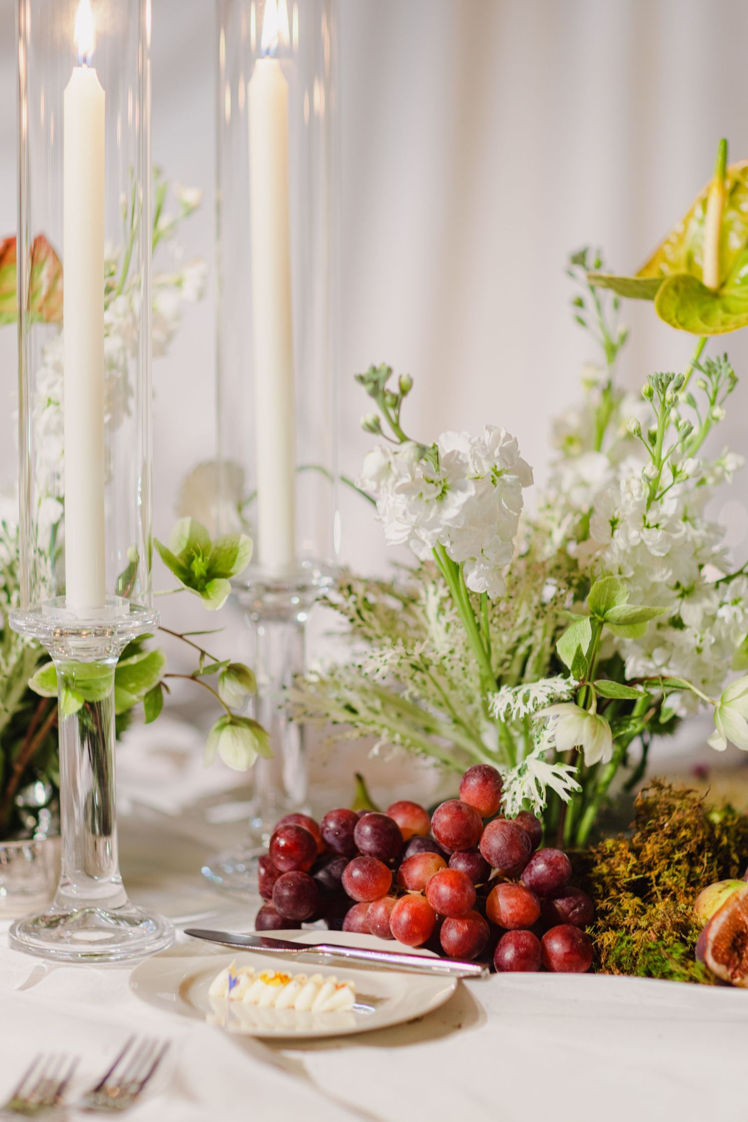Elegant table setting with white candles, fresh flowers, and red grapes on a white tablecloth at a formal dinner.