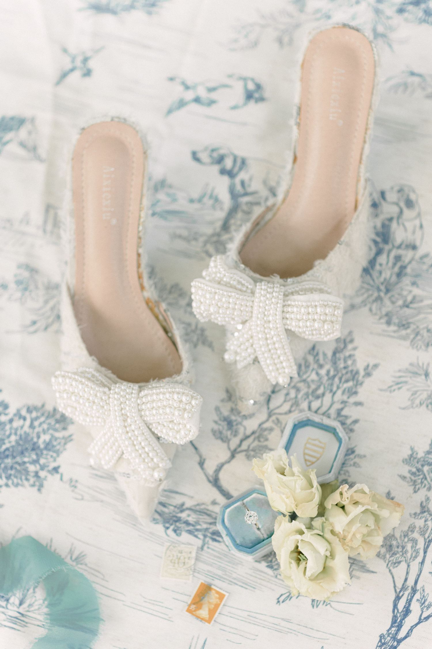 Pearl-embellished bridal shoes with bow details displayed on toile pattern background.