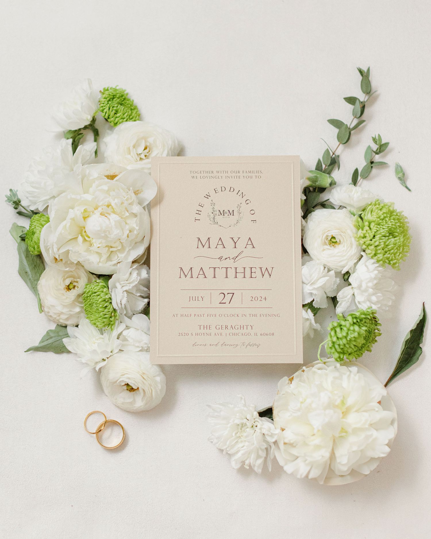 Wedding invitation with white and green florals and gold wedding bands on a white surface.