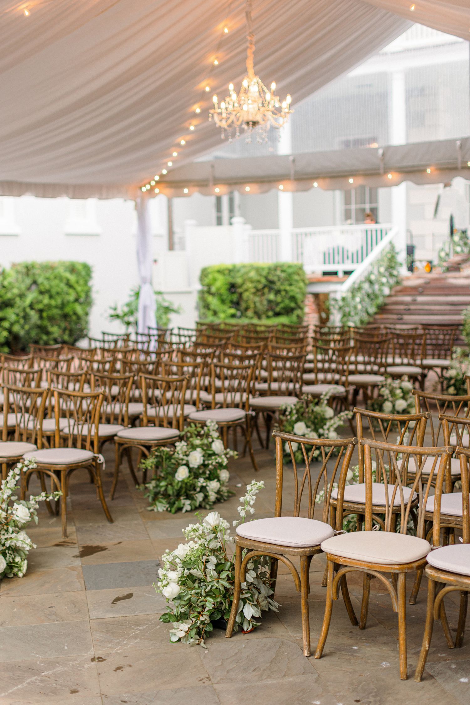 Elegant wedding ceremony setup with wooden chairs under white tent with chandelier and floral decorations.