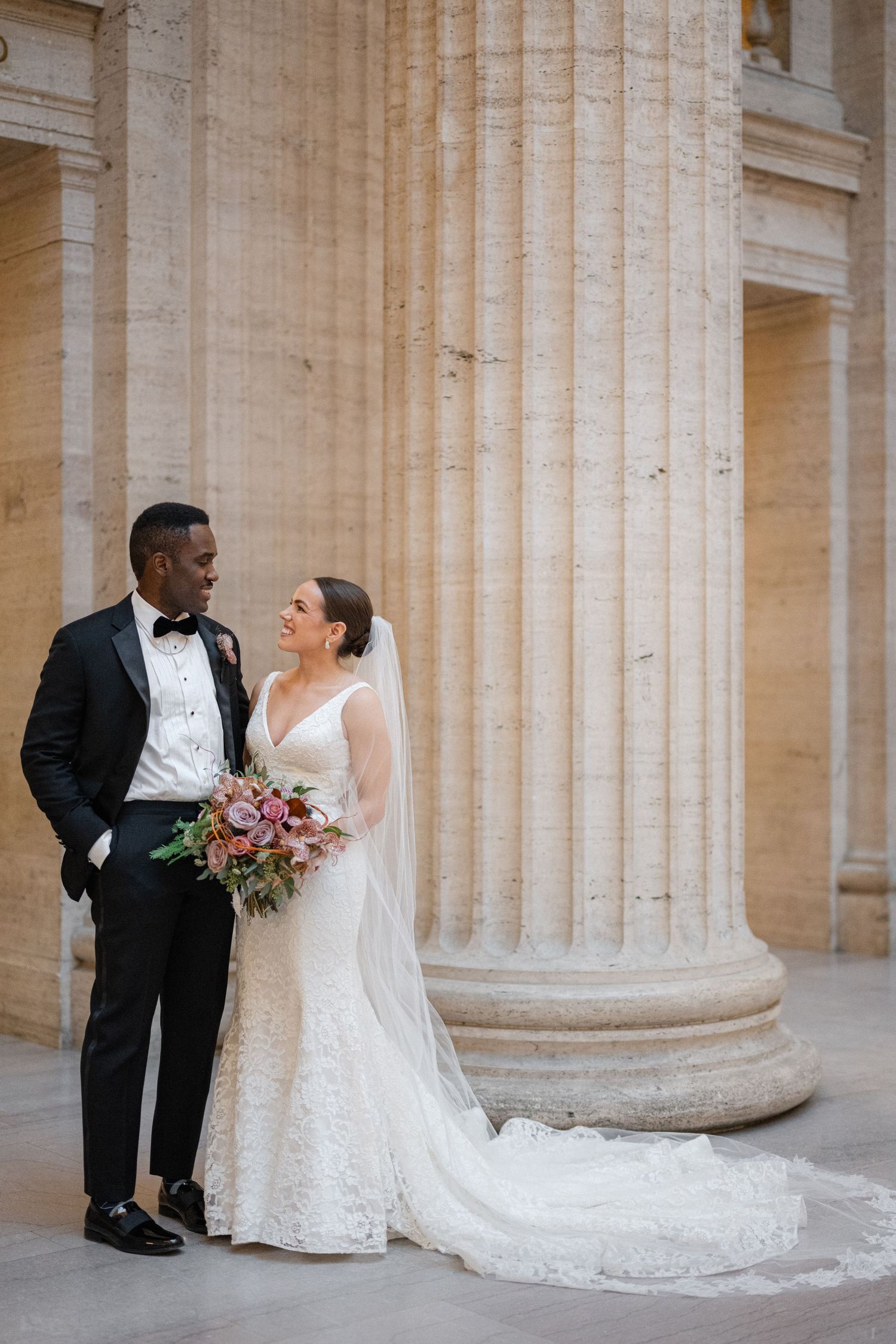 Romantic wedding portrait against grand classical stone columns with bride holding bouquet.