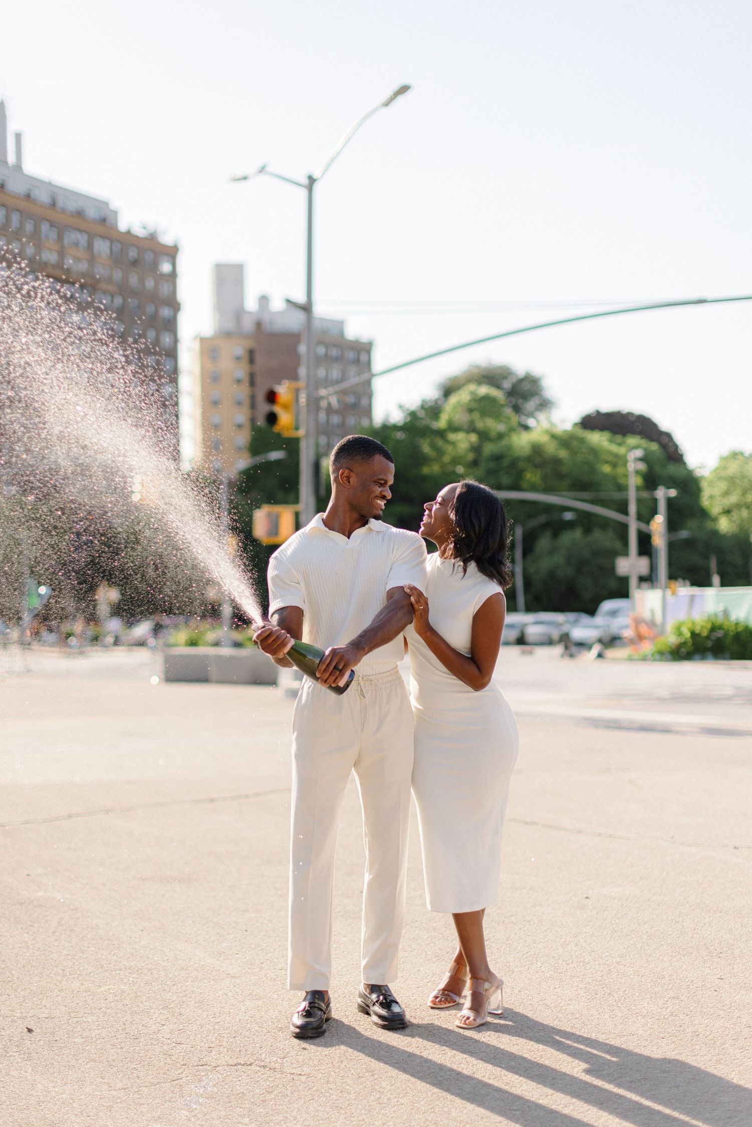 Playful couple in white outfits enjoying a spray of water on a sunny city street.