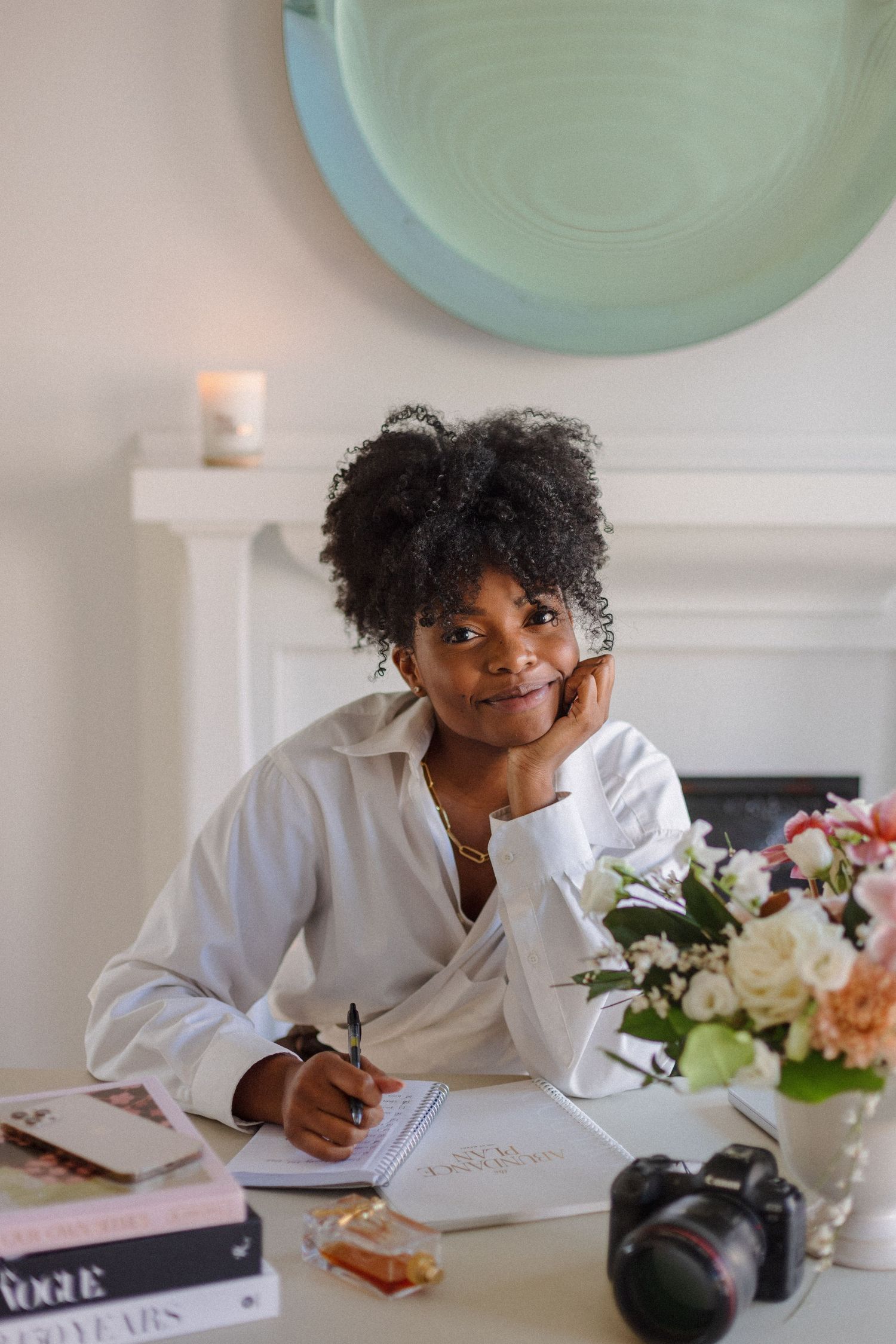 A person in a white shirt writes in a notebook at a desk decorated with flowers and decorative plates on the wall.
