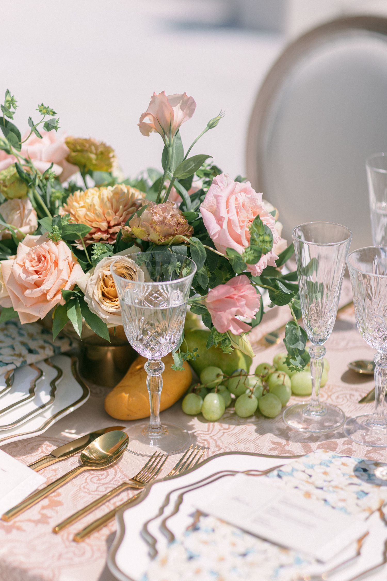 Romantic table setting with crystal glasses, gold flatware, and peach floral centerpiece with fresh grapes.