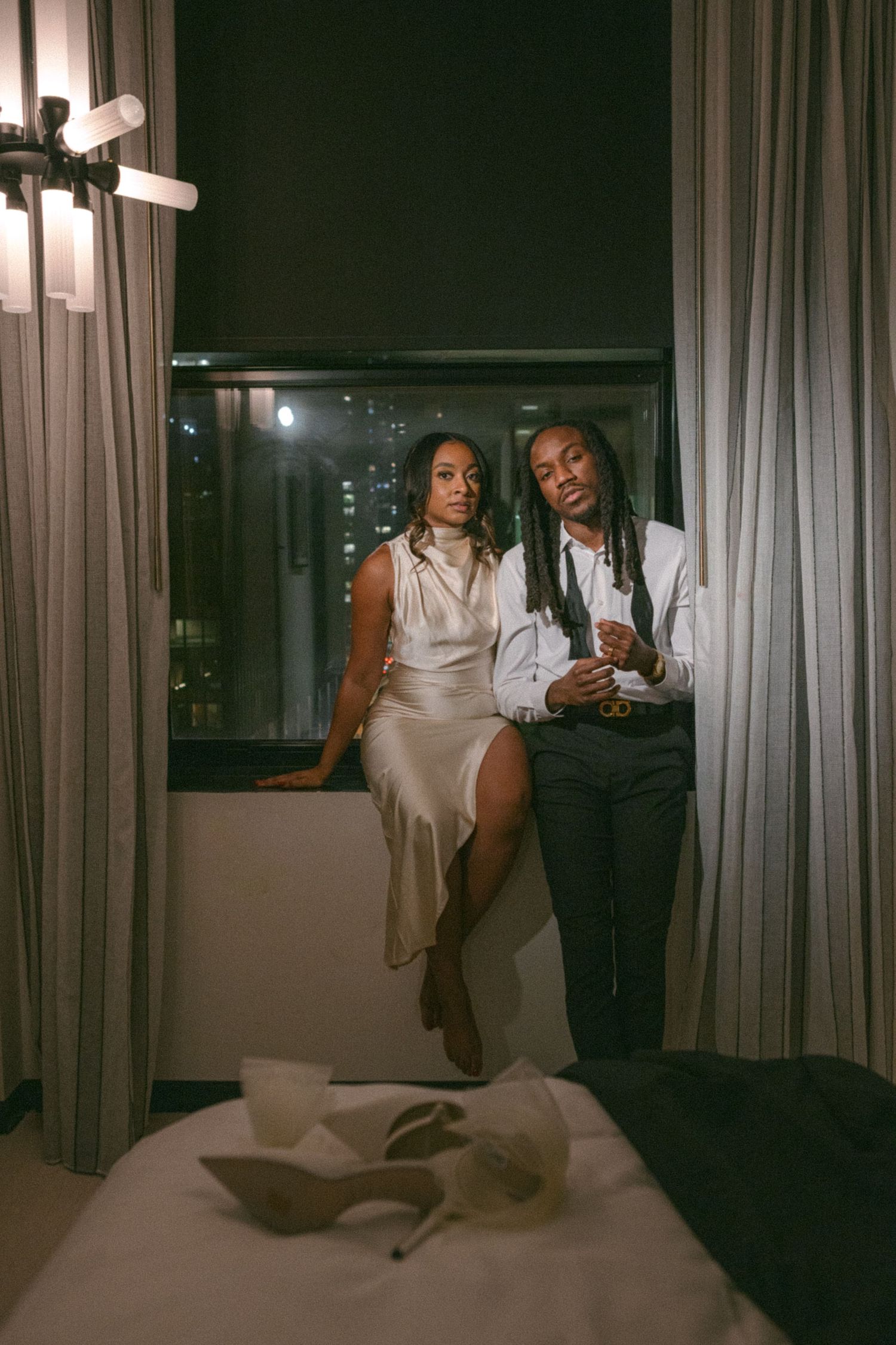 Two people in formal attire pose by a window in an evening cityscape.