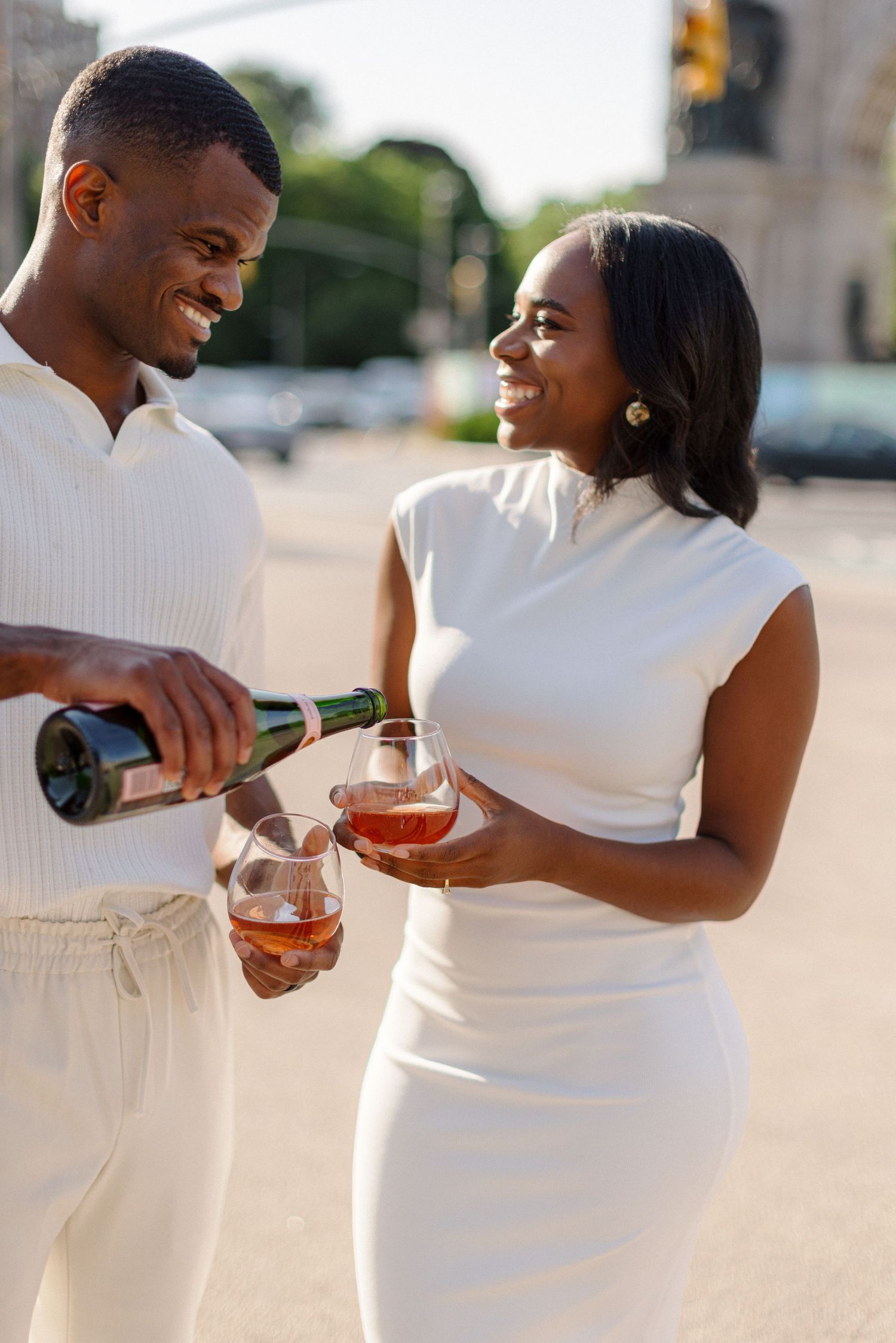 A couple in white outfits pouring and sharing wine outdoors on a sunny day.