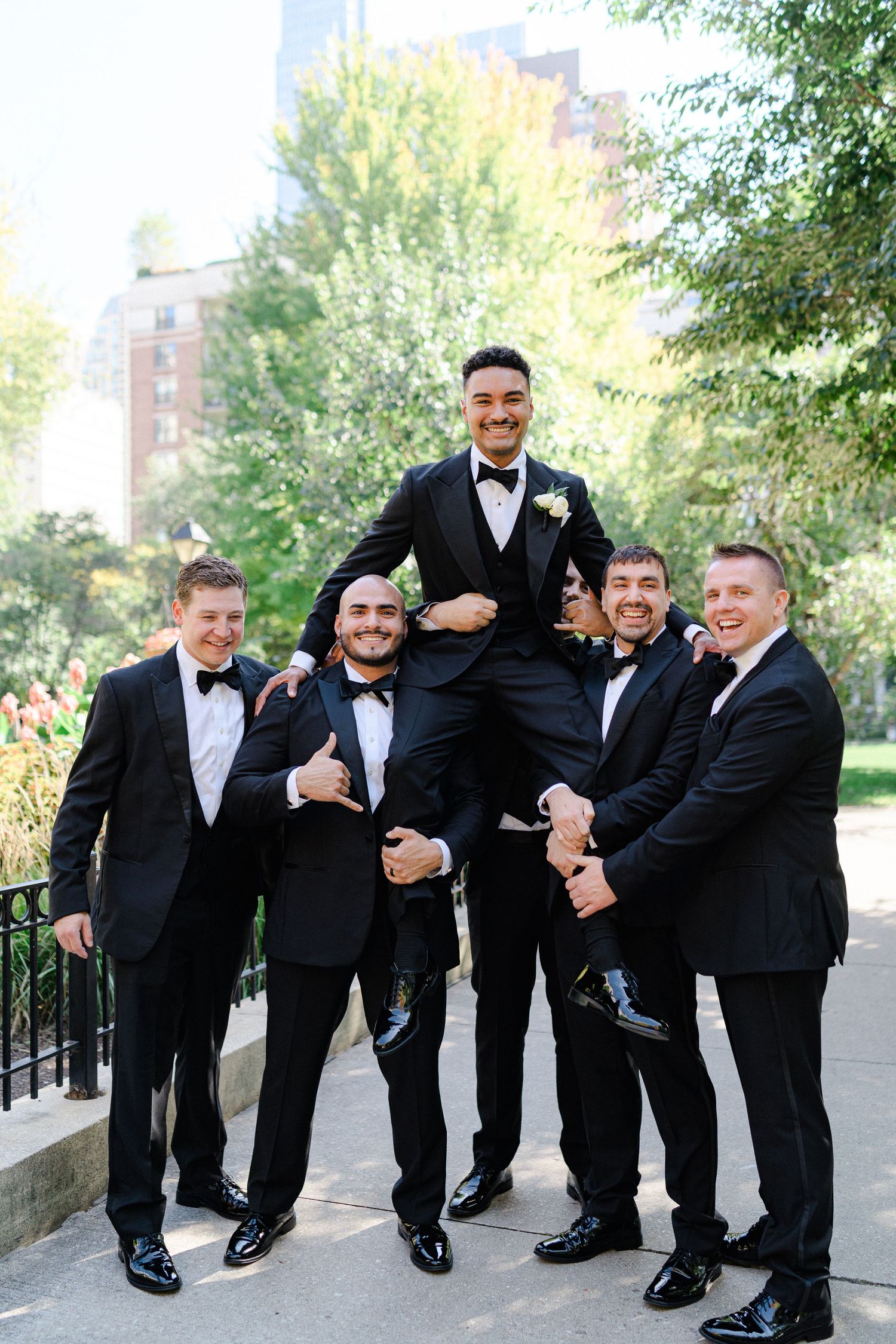 Groomsmen in black tuxedos lift and support one member of the wedding party in a celebratory pose.