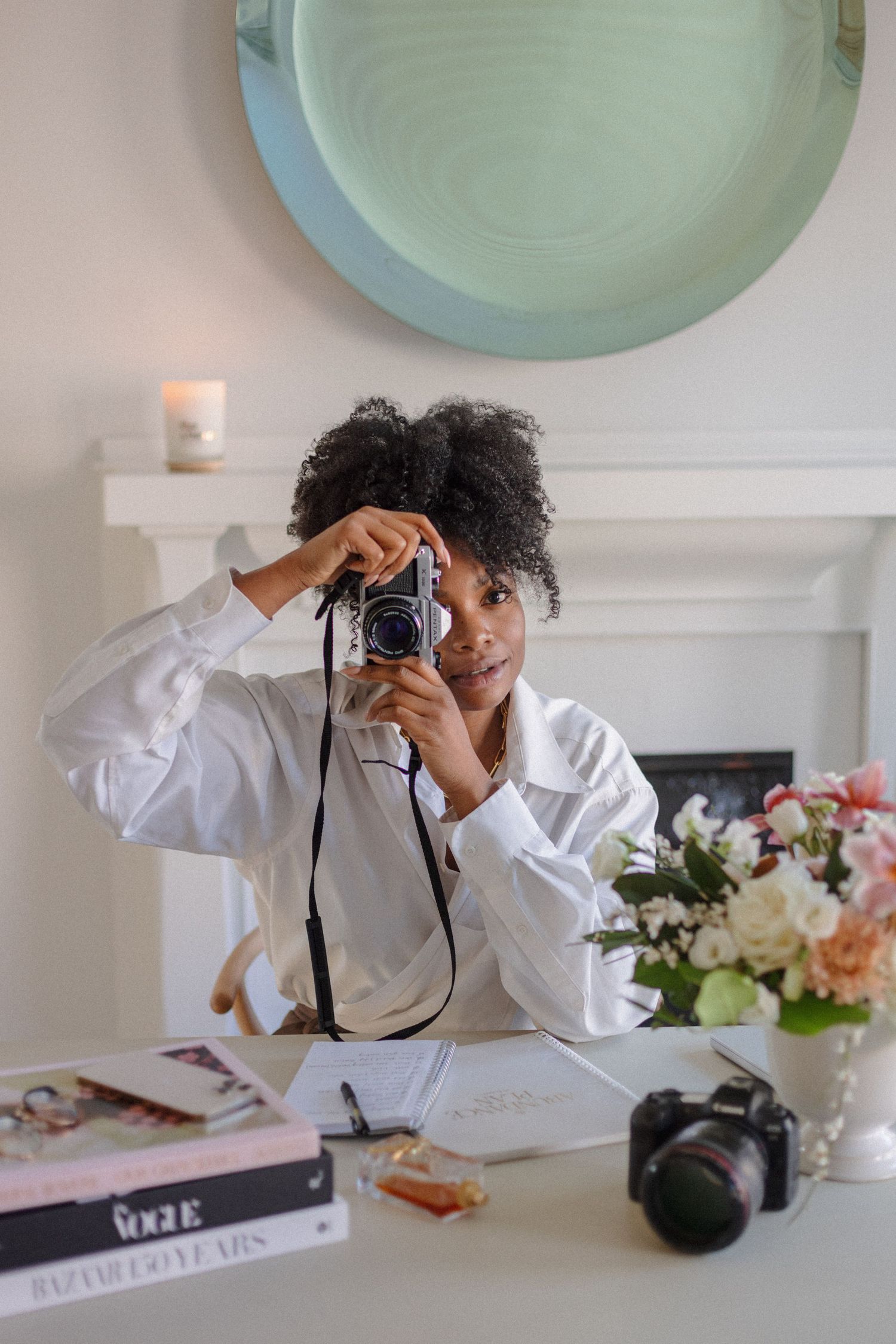 Photographer using vintage camera at desk with books and floral arrangements.
