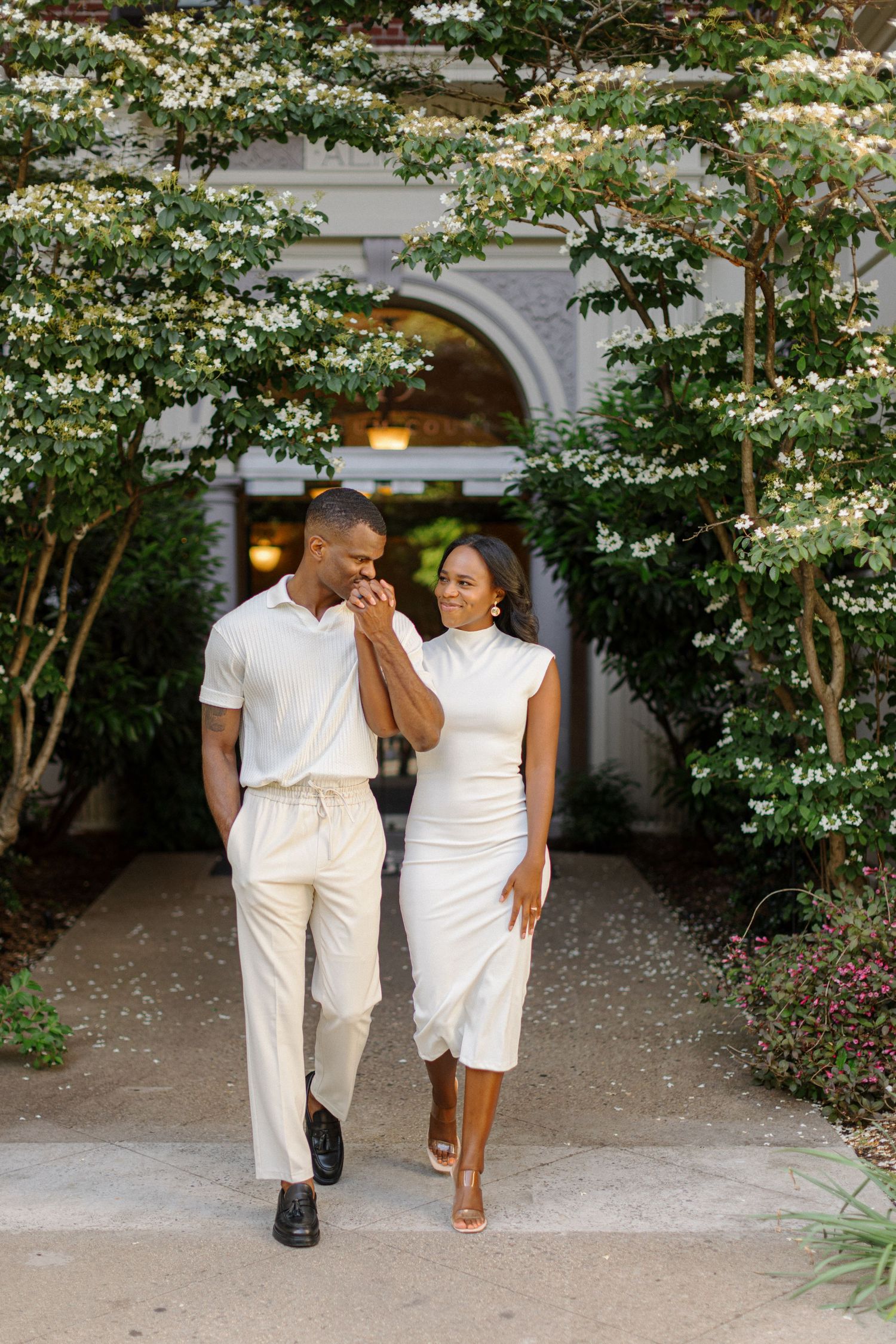 Couple in matching white outfits walking together through a flowering garden archway.