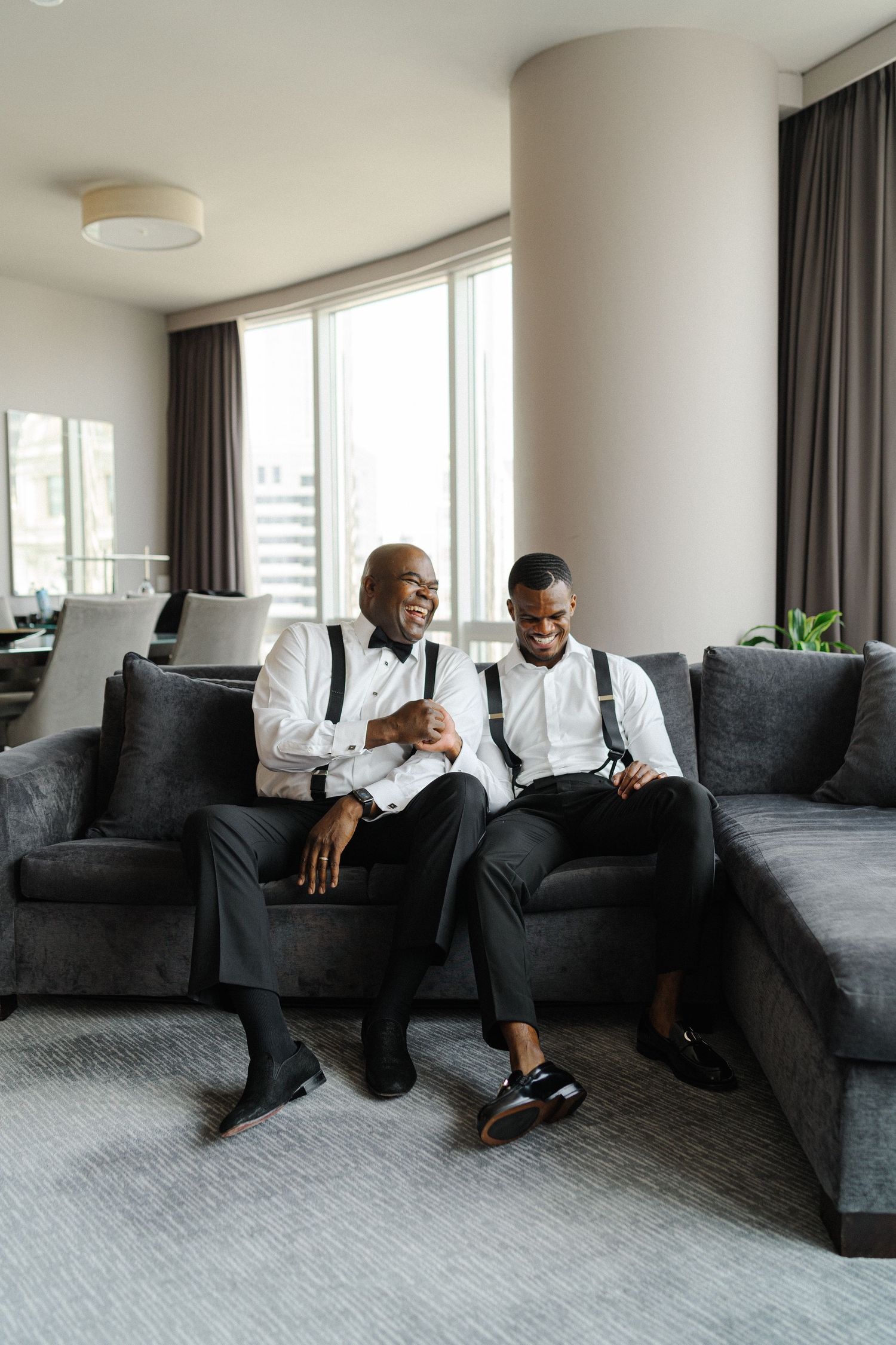Two people in formal attire sharing a laugh while relaxing on a modern gray couch.
