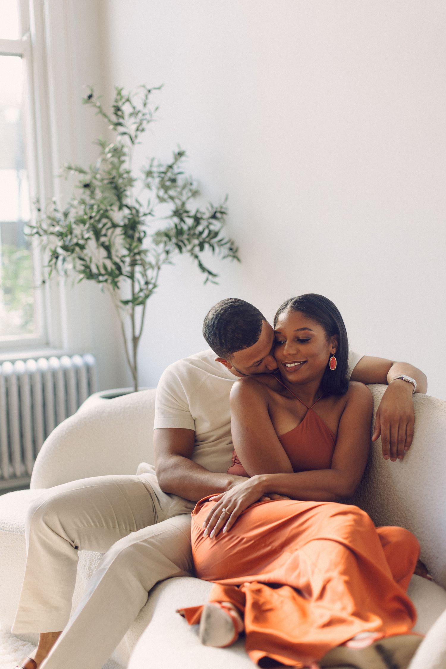 A couple shares an intimate moment while relaxing together on a white couch near a potted plant by a window.