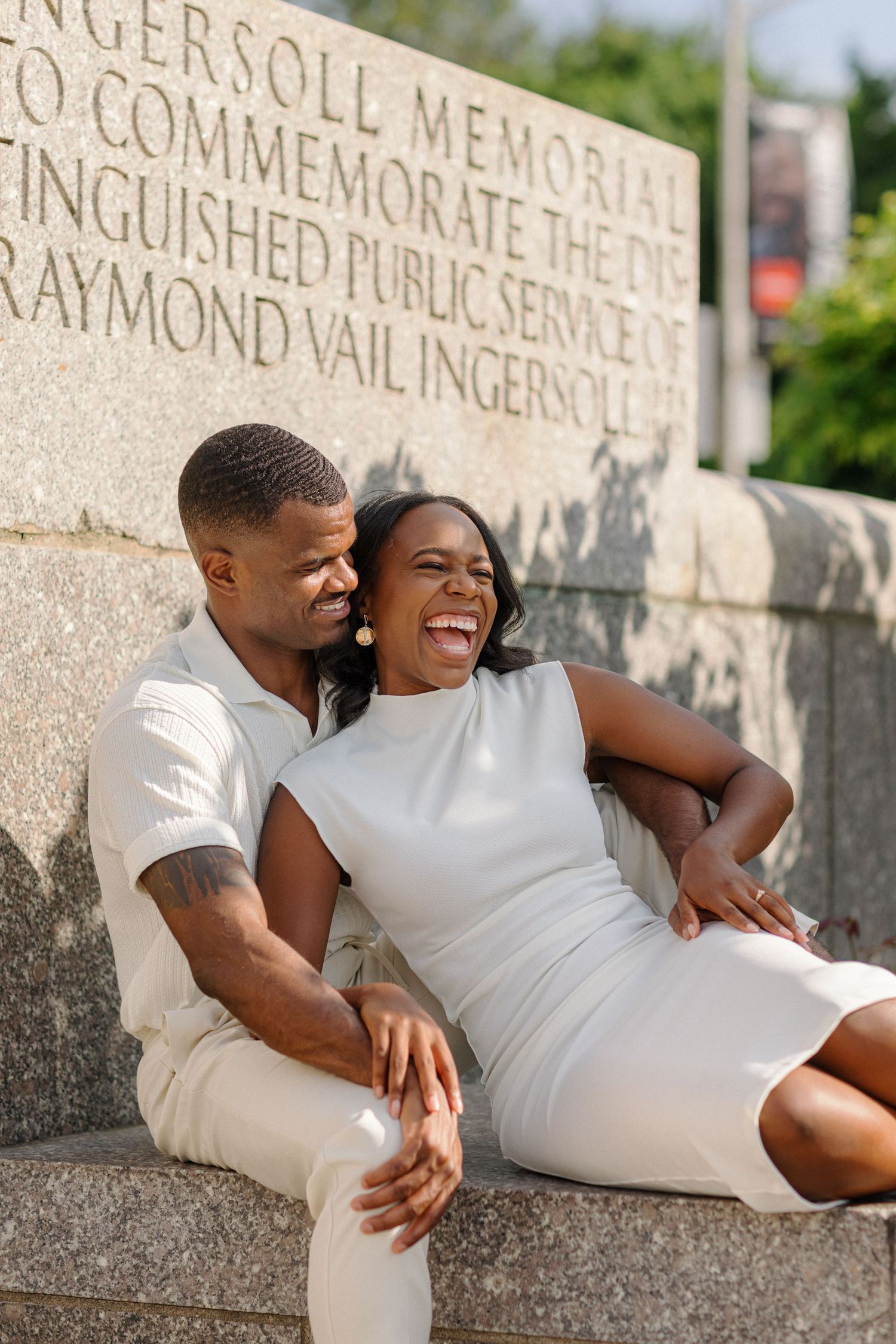 Happy couple in white attire share a joyful moment while relaxing on stone steps near a memorial.