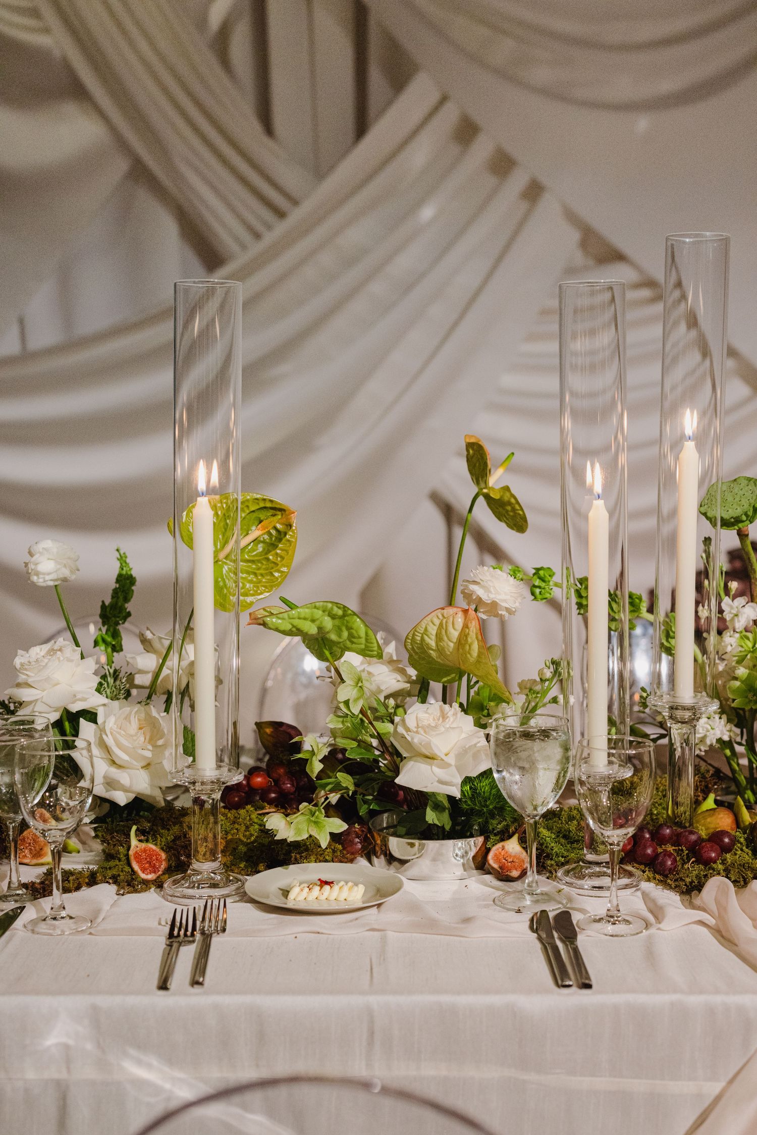 Elegant wedding table setting with white roses, figs, and tall glass candleholders against draped fabric backdrop.
