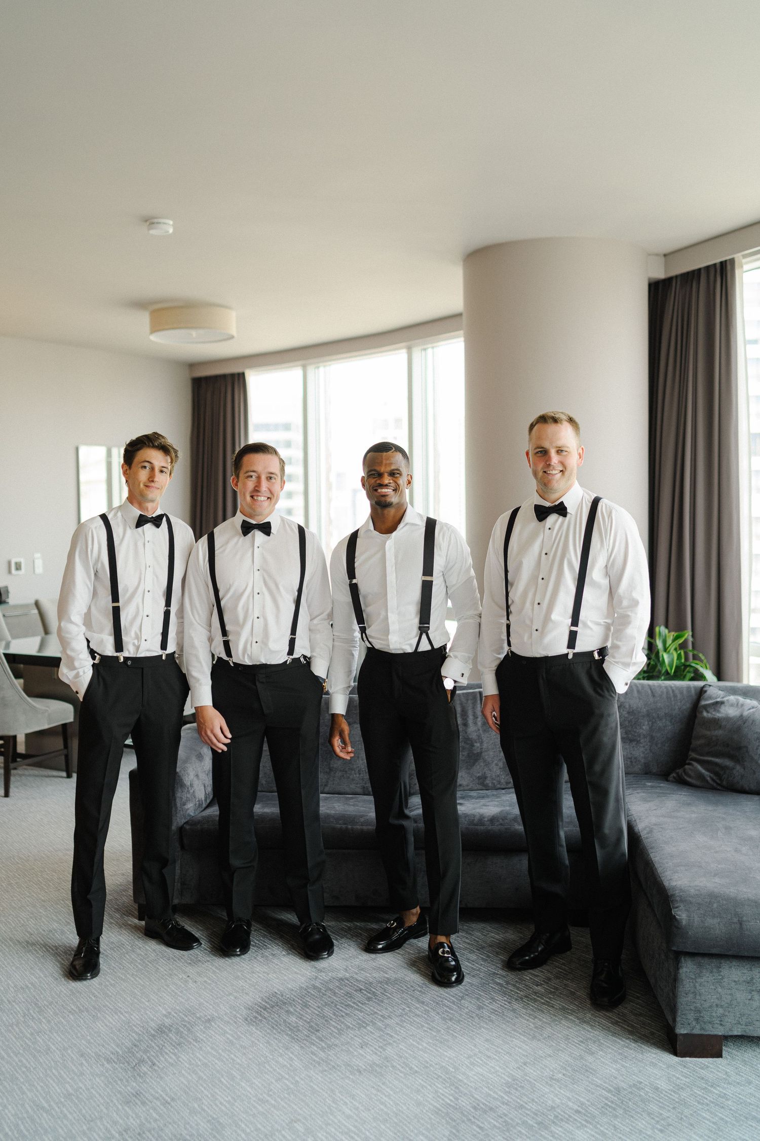 Four people wearing matching black bow ties and suspenders pose together in front of gray couch.