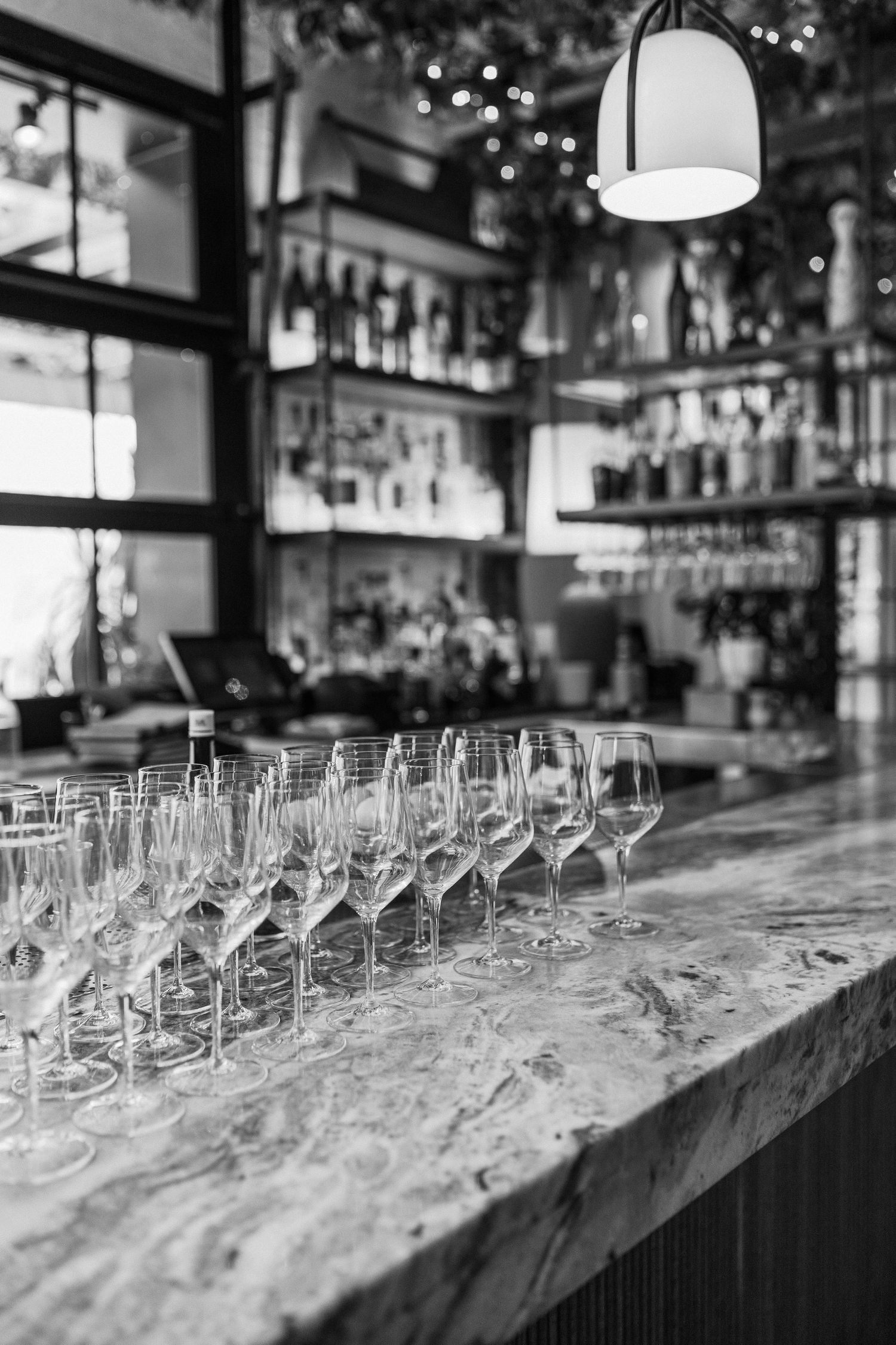 Black and white photo of wine glasses lined up on marble bar counter with bottle display in background.