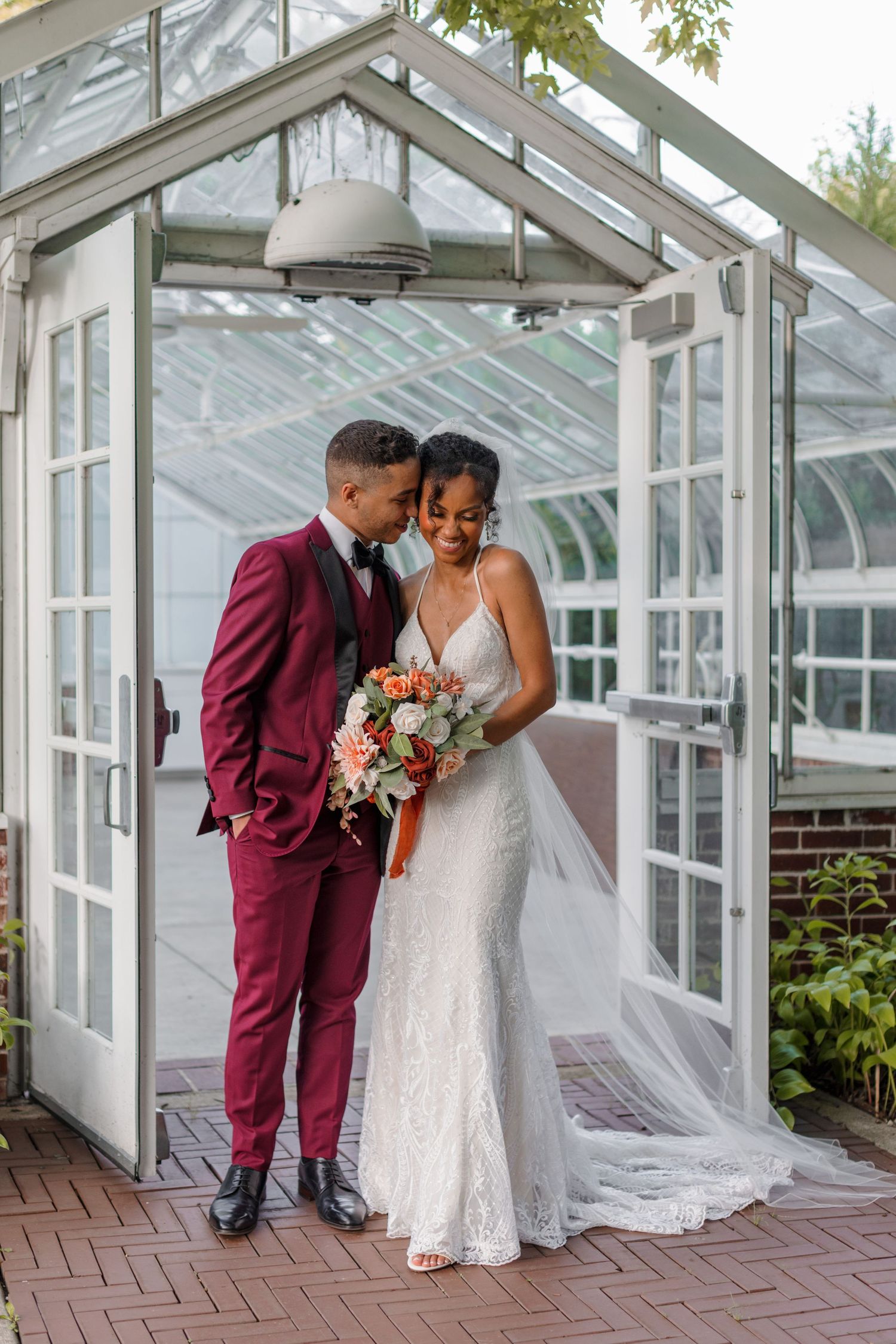 A romantic greenhouse wedding photo featuring a couple in burgundy suit and white dress with floral bouquet.