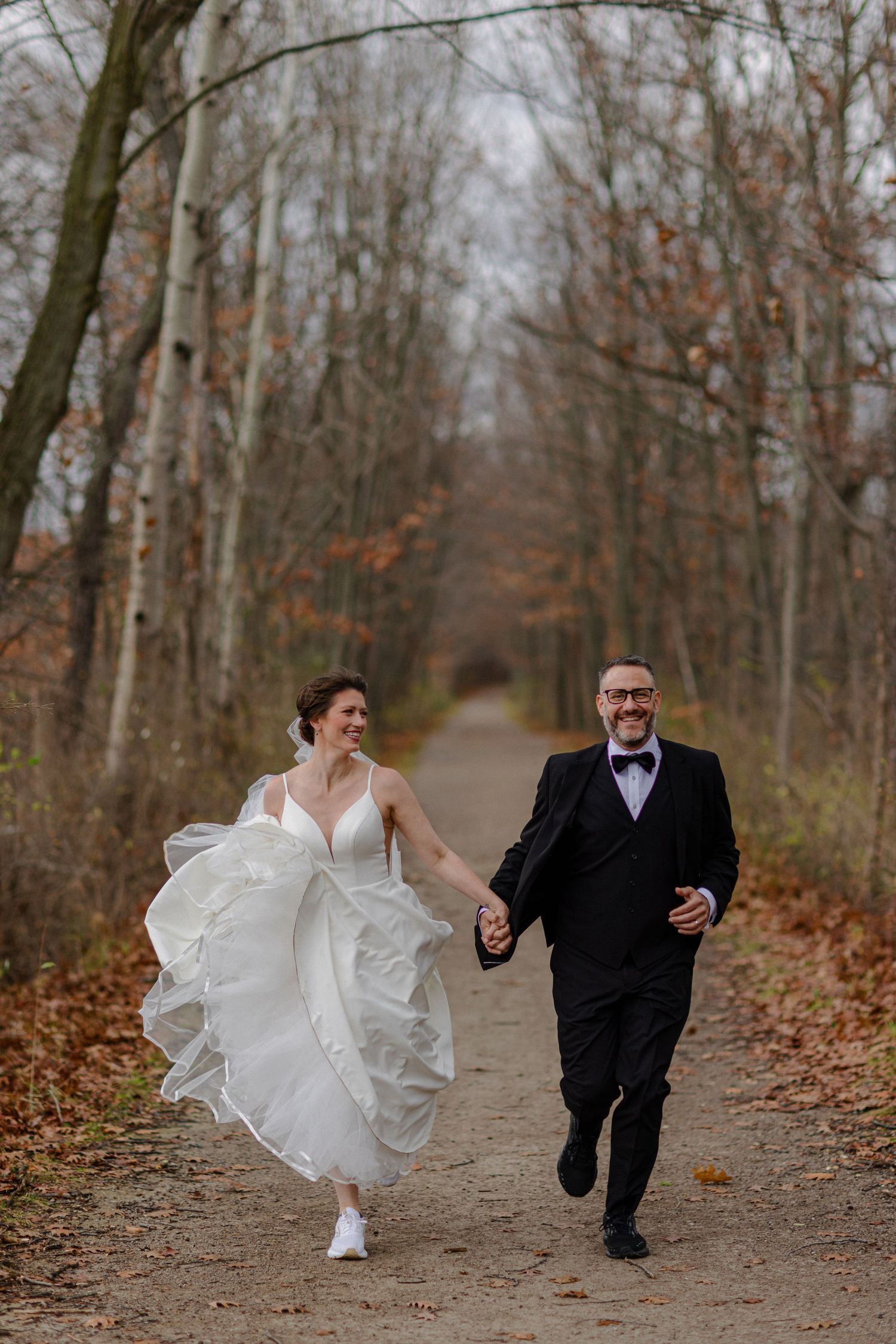 A joyful couple running hand-in-hand down an autumn forest path in wedding attire.