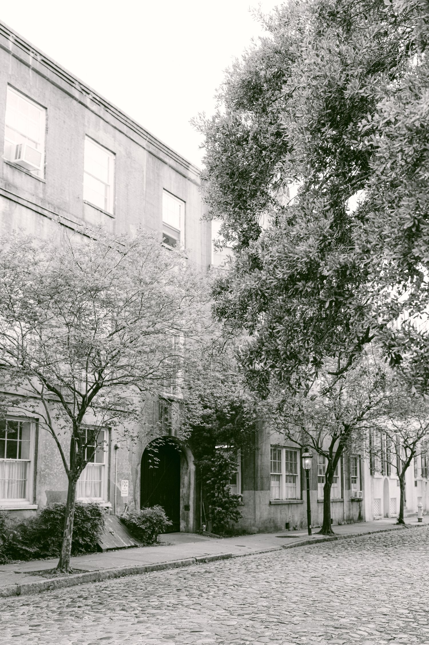 Black and white photograph of a historic building with arched entrance and trees along cobblestone street.