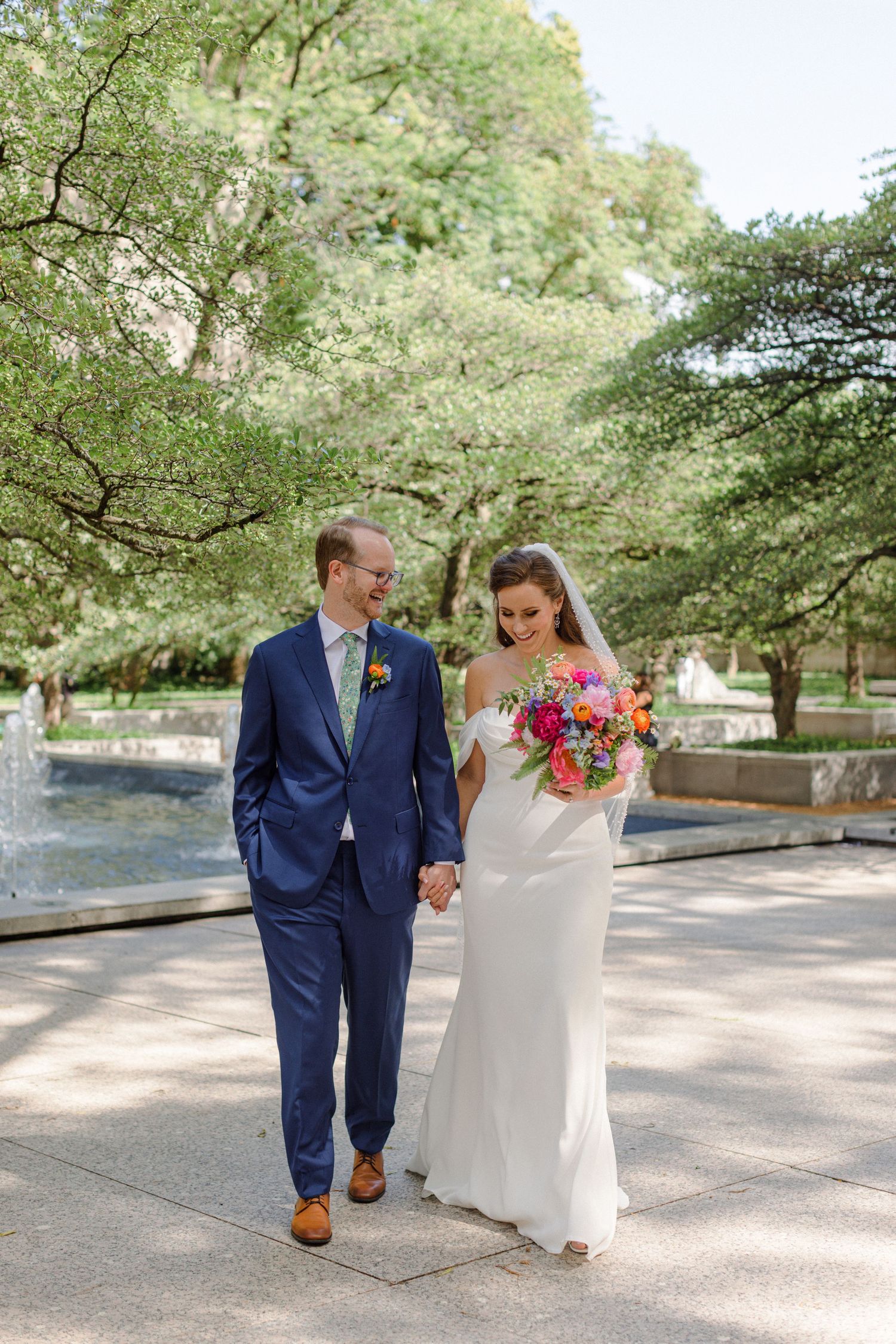 A couple walks together outdoors, with the bride holding a vibrant pink bouquet against a backdrop of trees and fountain.